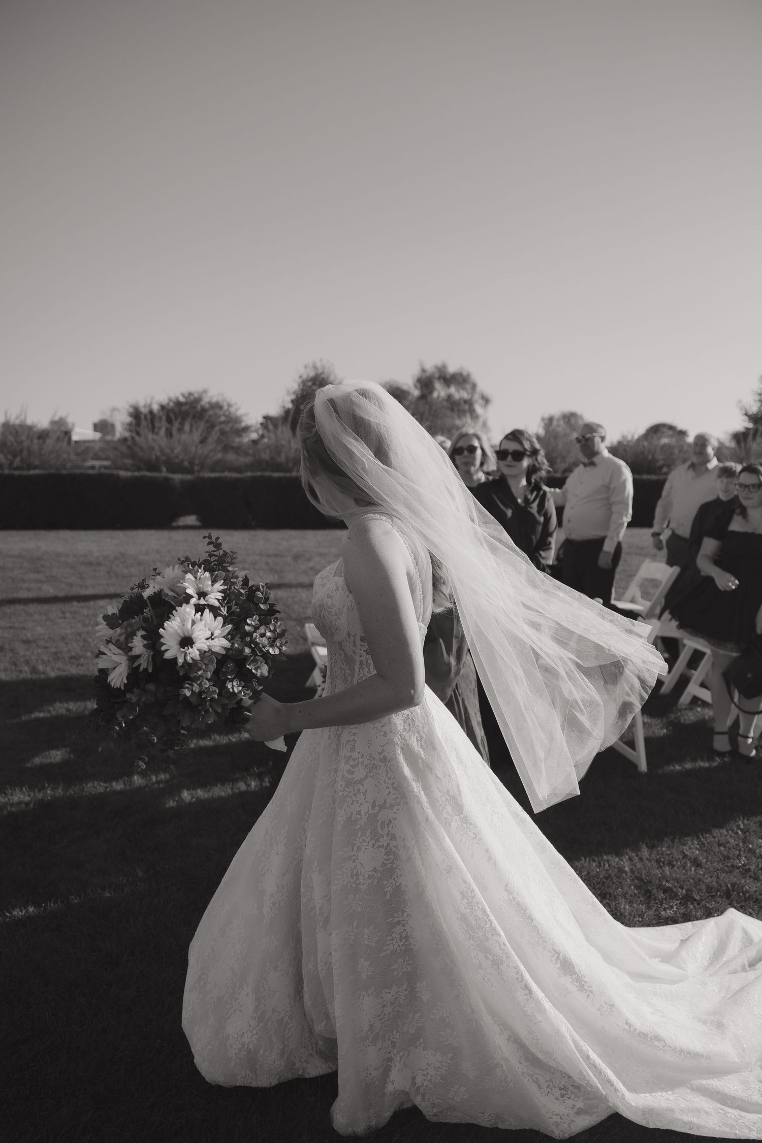 Black and white photo of a bride in lace gown and veil holding bouquet at outdoor wedding ceremony.
