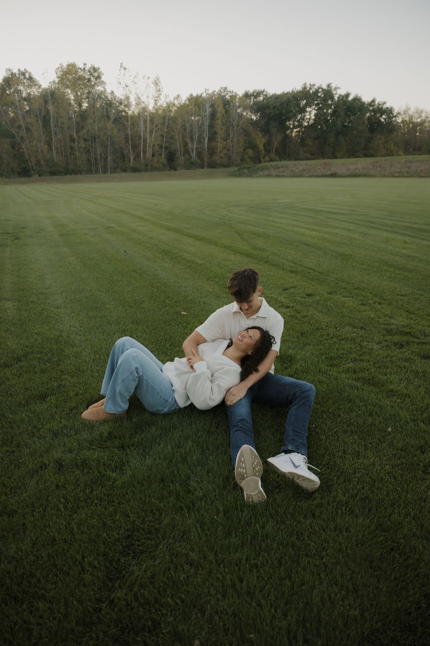 A couple relaxes together on a freshly mowed green lawn at sunset with trees in the background.