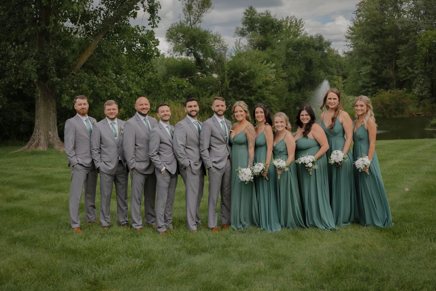 Wedding party lined up outdoors wearing coordinating emerald green dresses and gray suits.