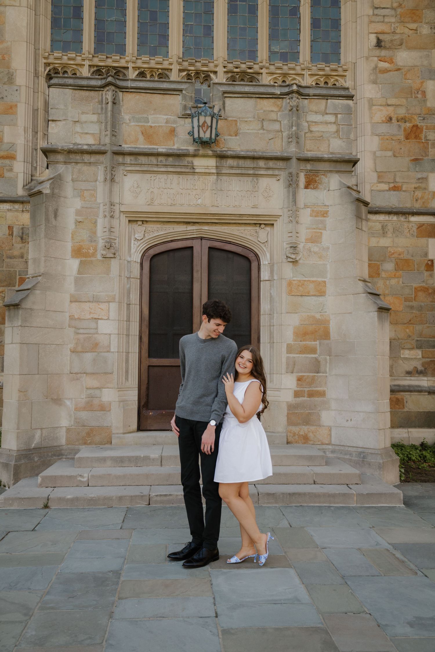 A couple poses romantically in front of a historic stone building, woman in white dress, man in gray sweater.