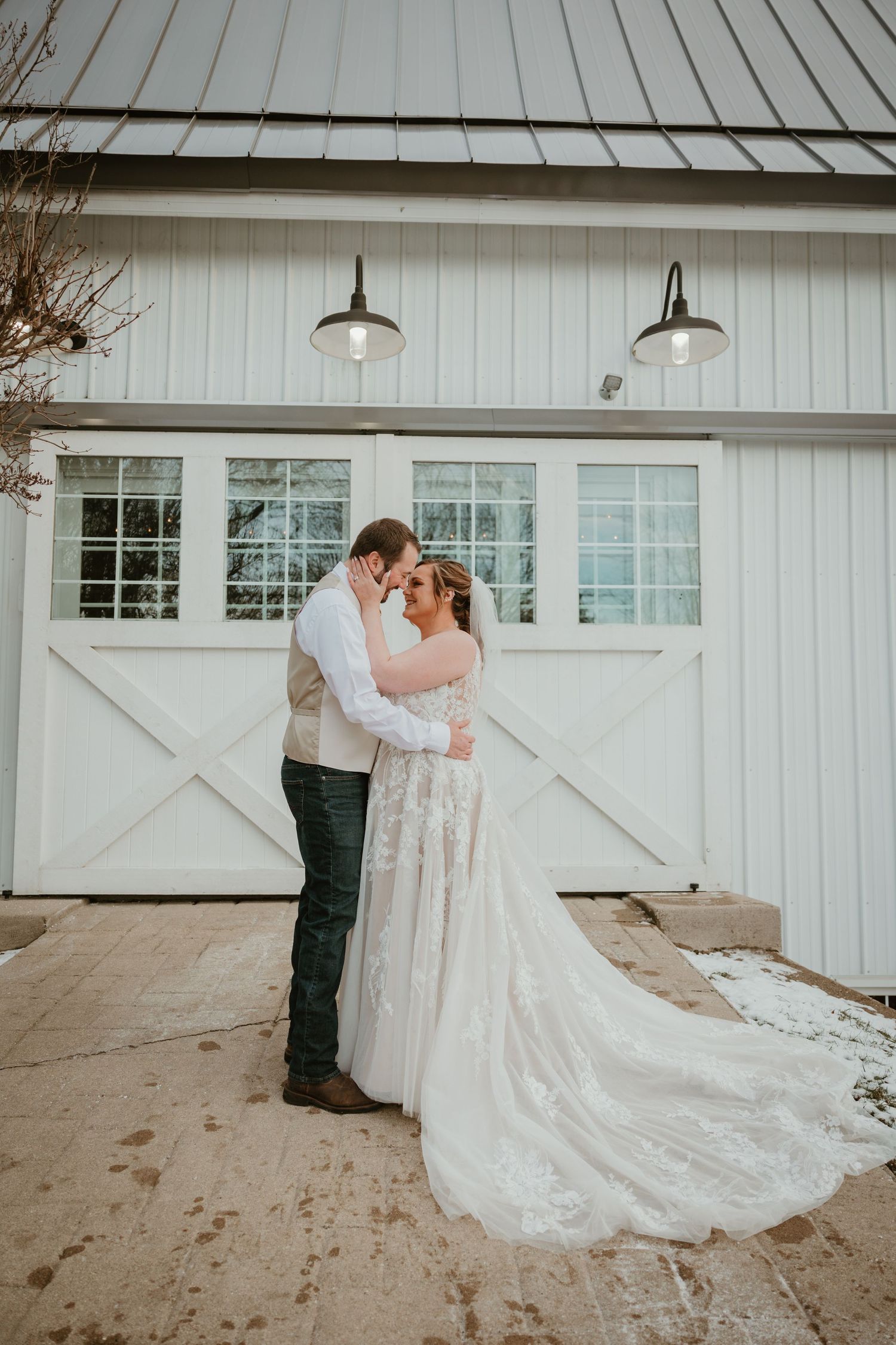 A romantic wedding portrait of a couple embracing in front of a white barn with pendant lights.