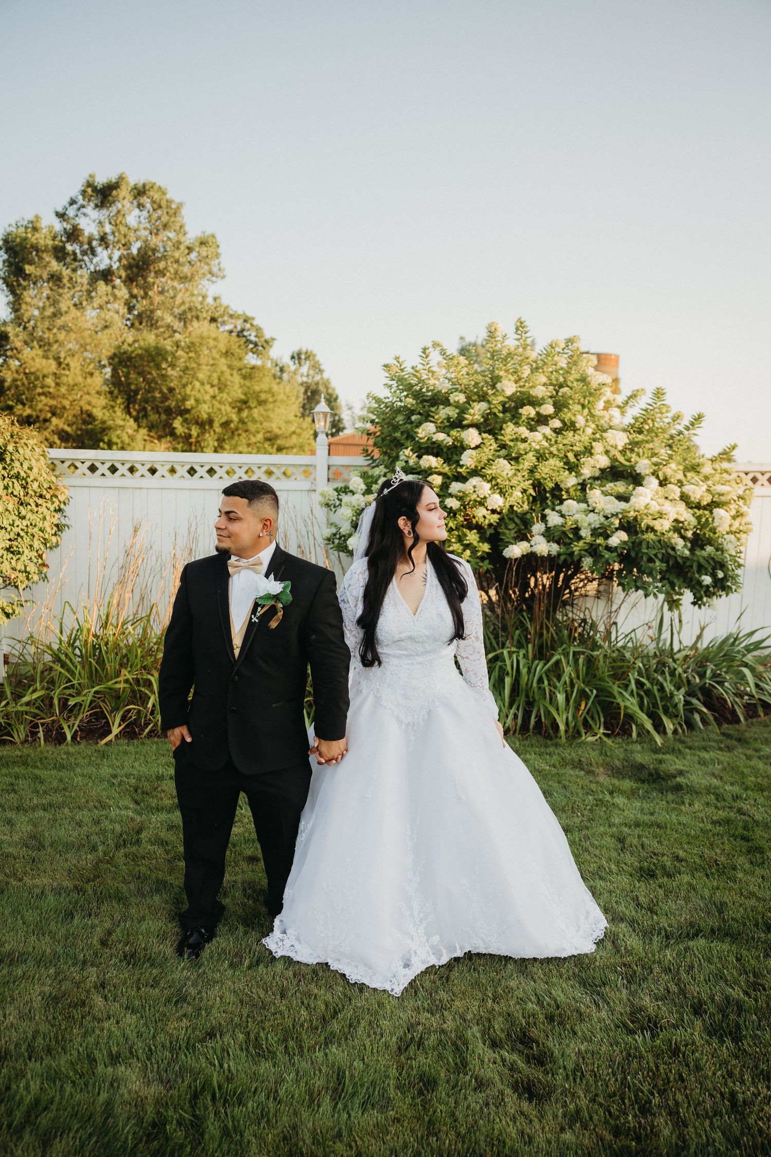 A couple in wedding attire walking through a garden with white fence and greenery.