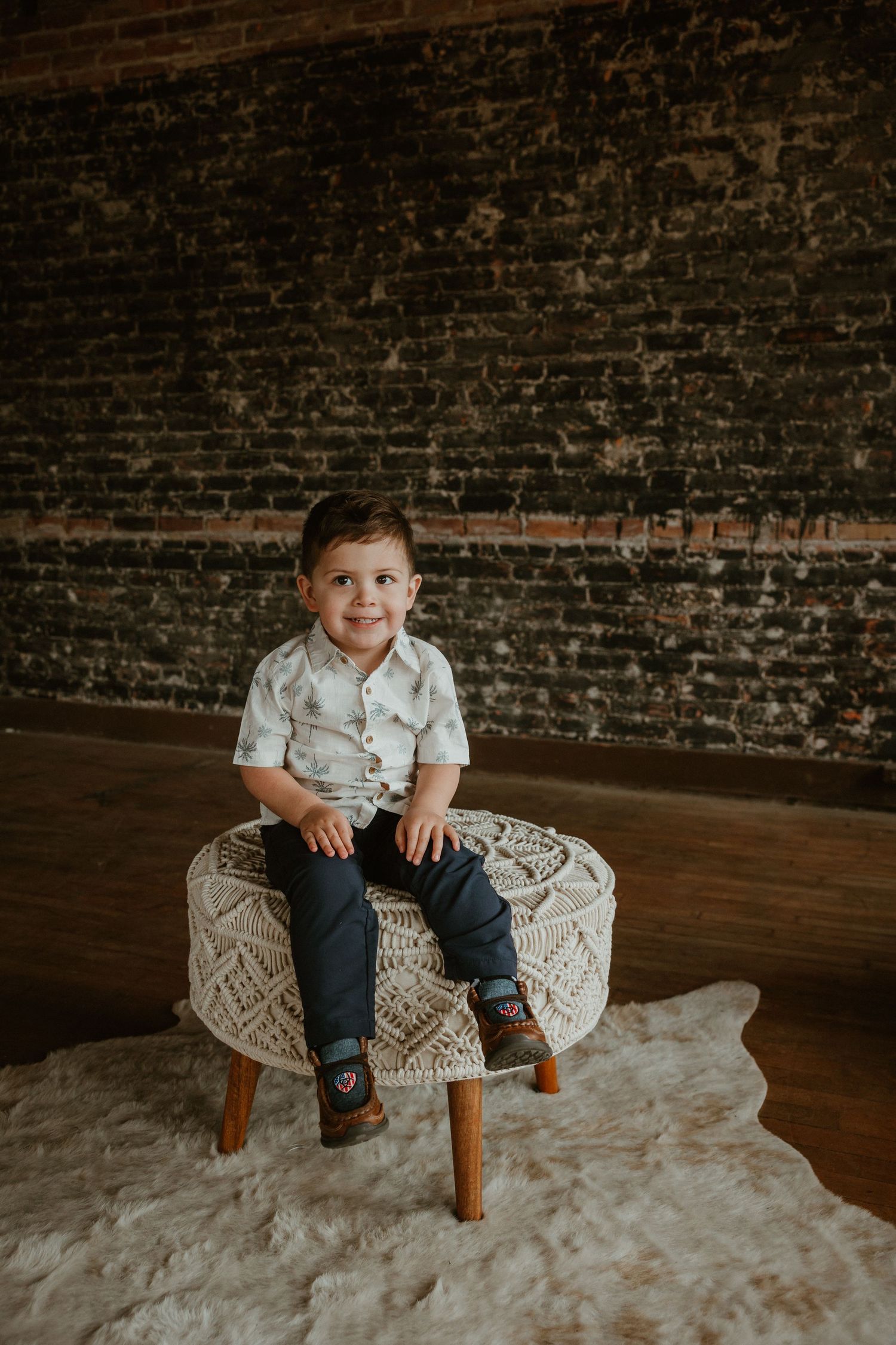 Young boy in white shirt sitting on a macrame ottoman against rustic brick wall background.