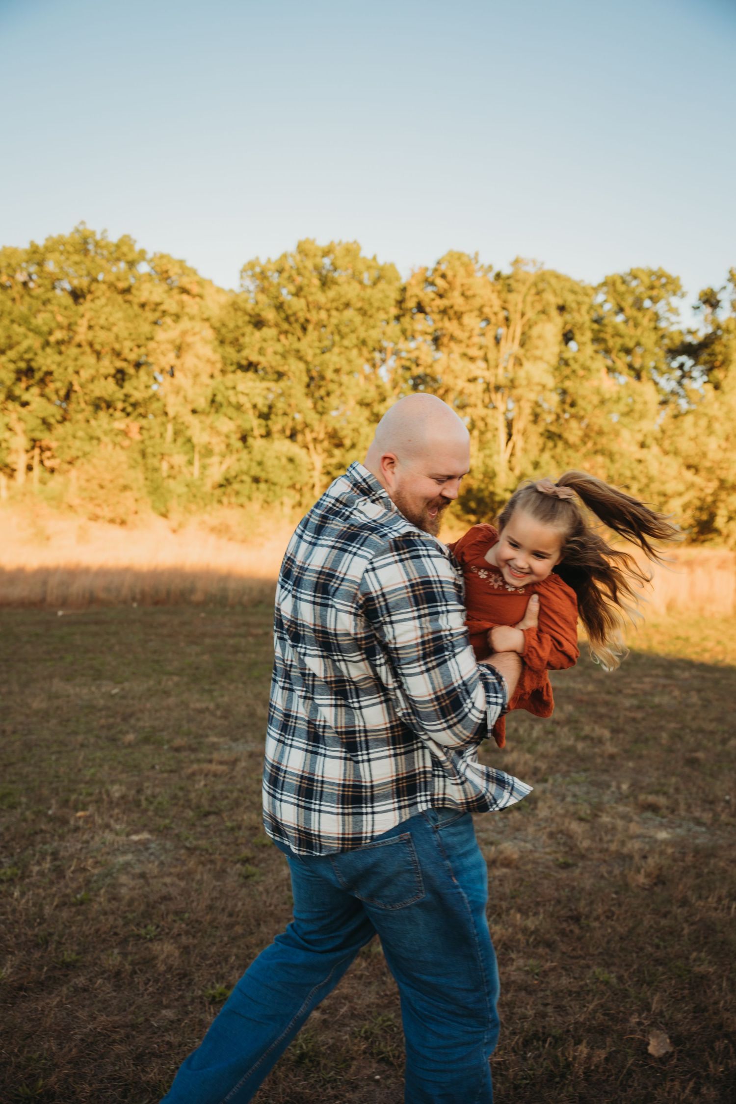 Adult in plaid shirt playfully swinging child in orange dress in outdoor field setting.