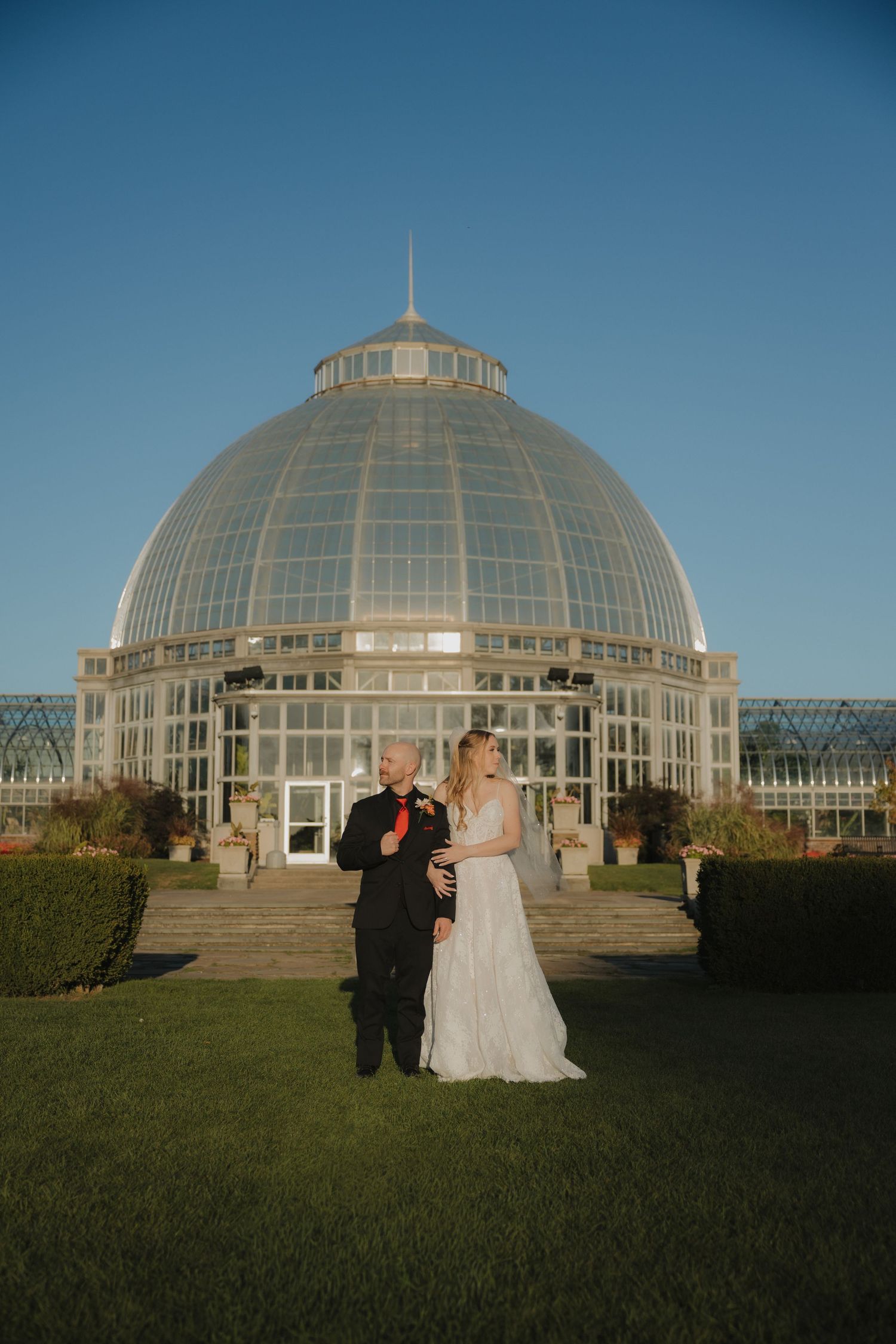 Bride and groom stand together on lawn before a stunning glass dome conservatory venue.