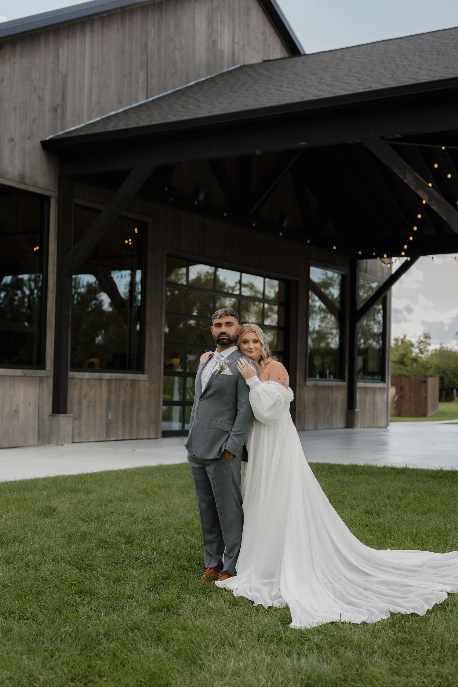 A couple in wedding attire embraces on a grassy lawn outside a modern barn-style venue with string lights.