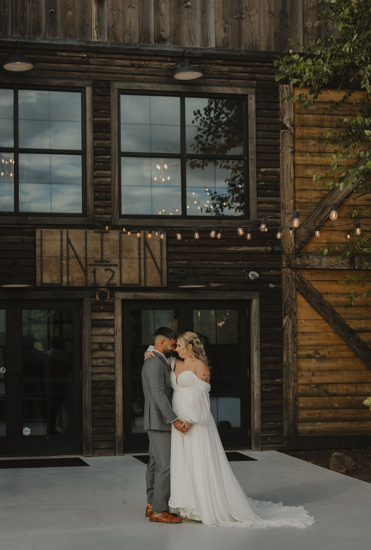 Wedding couple poses against rustic wooden venue with string lights and large windows.