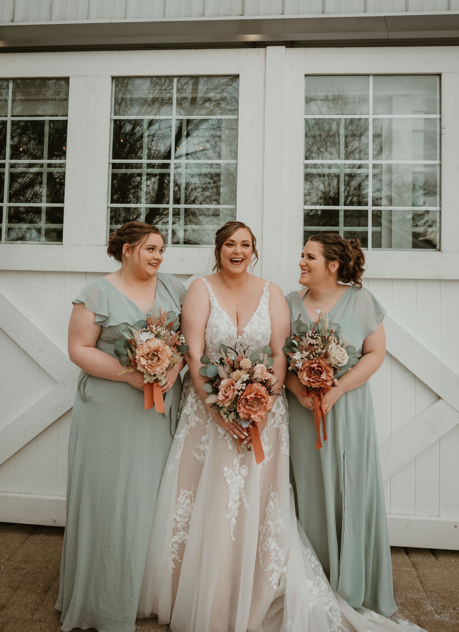 Three women in sage green and blush dresses pose with bouquets against a white barn backdrop.