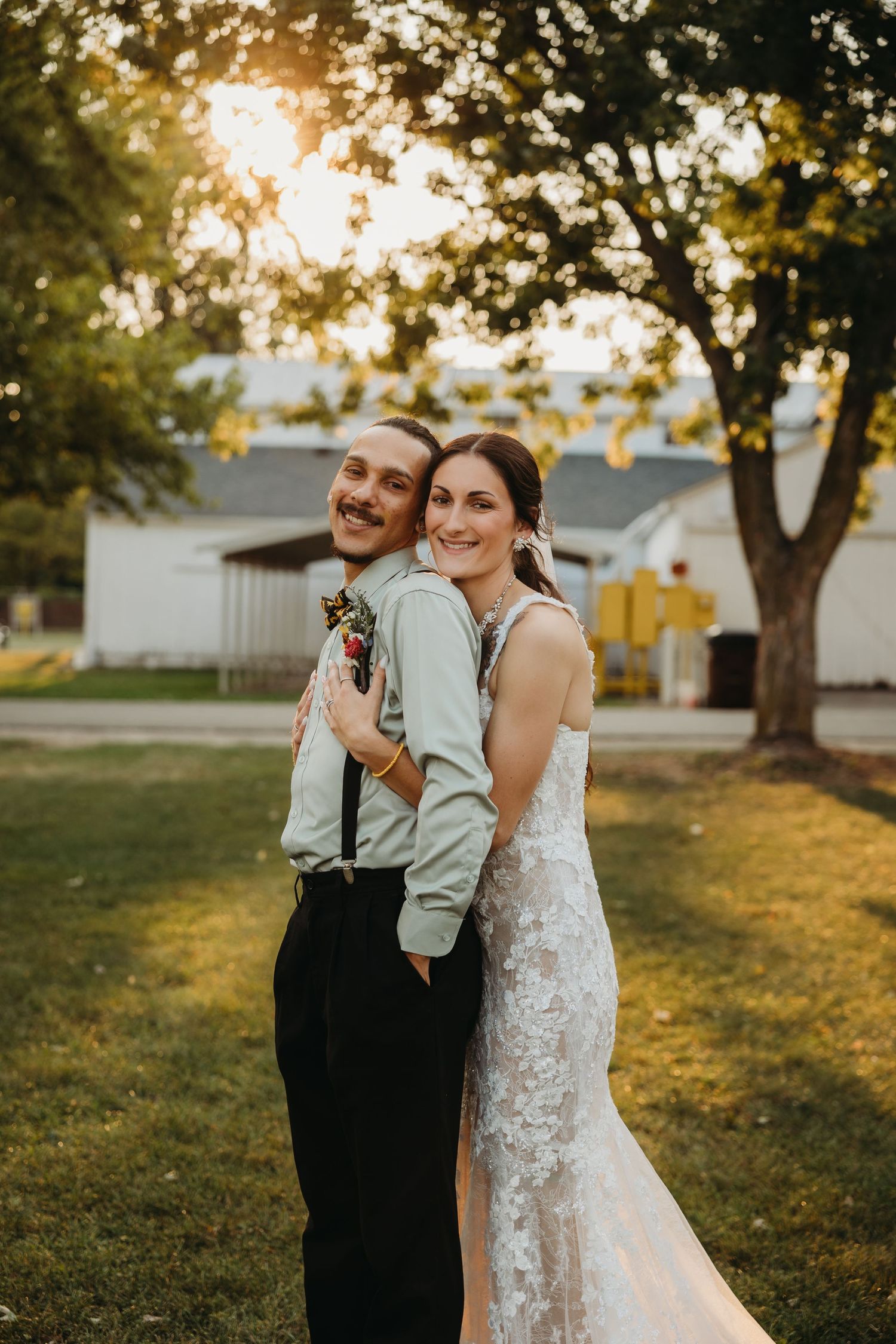 Bride in lace dress and groom in suspenders share a candid moment at sunset in a backyard garden setting.