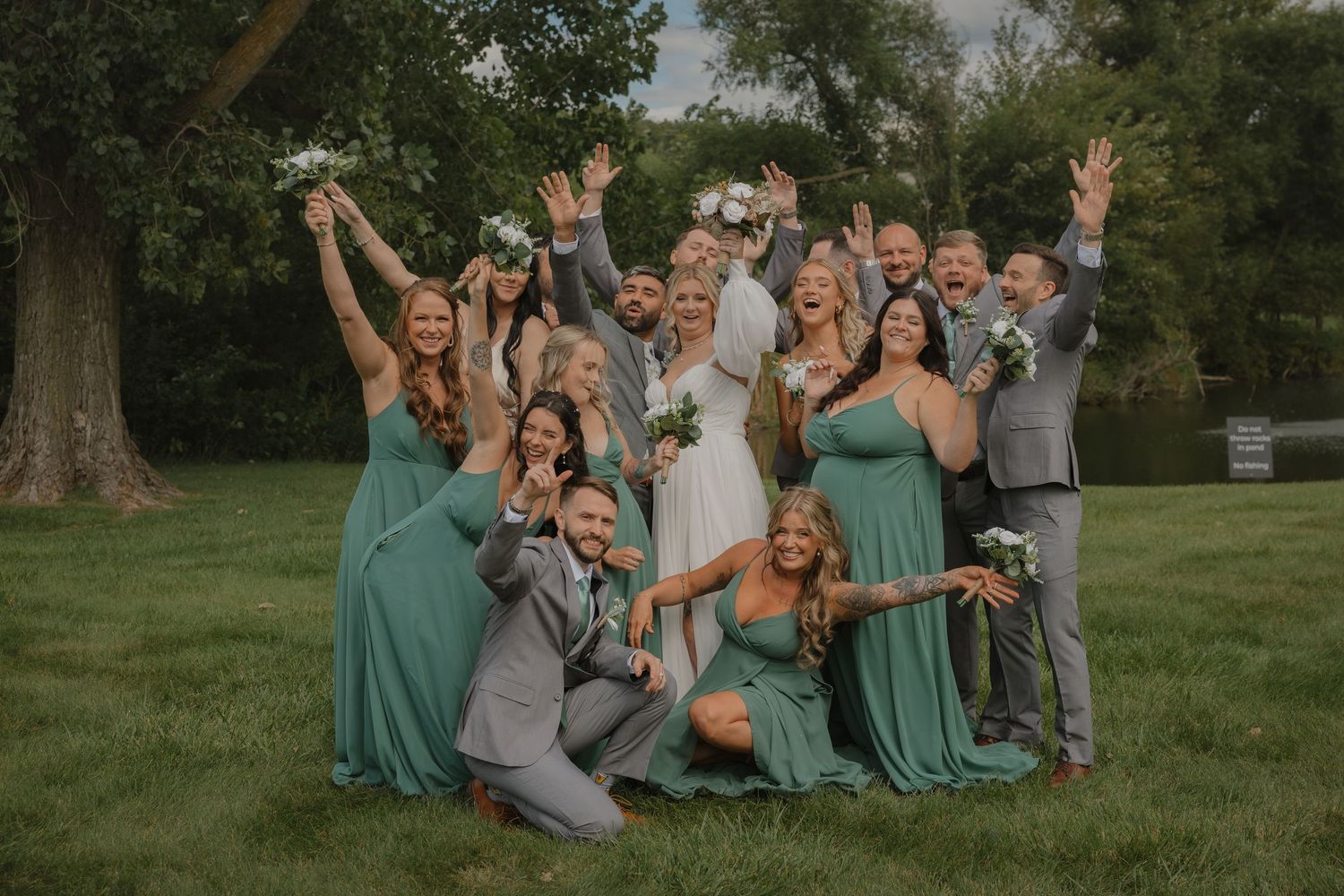 Wedding party celebrates joyfully on grass with bridesmaids in green dresses and groomsmen in gray suits.