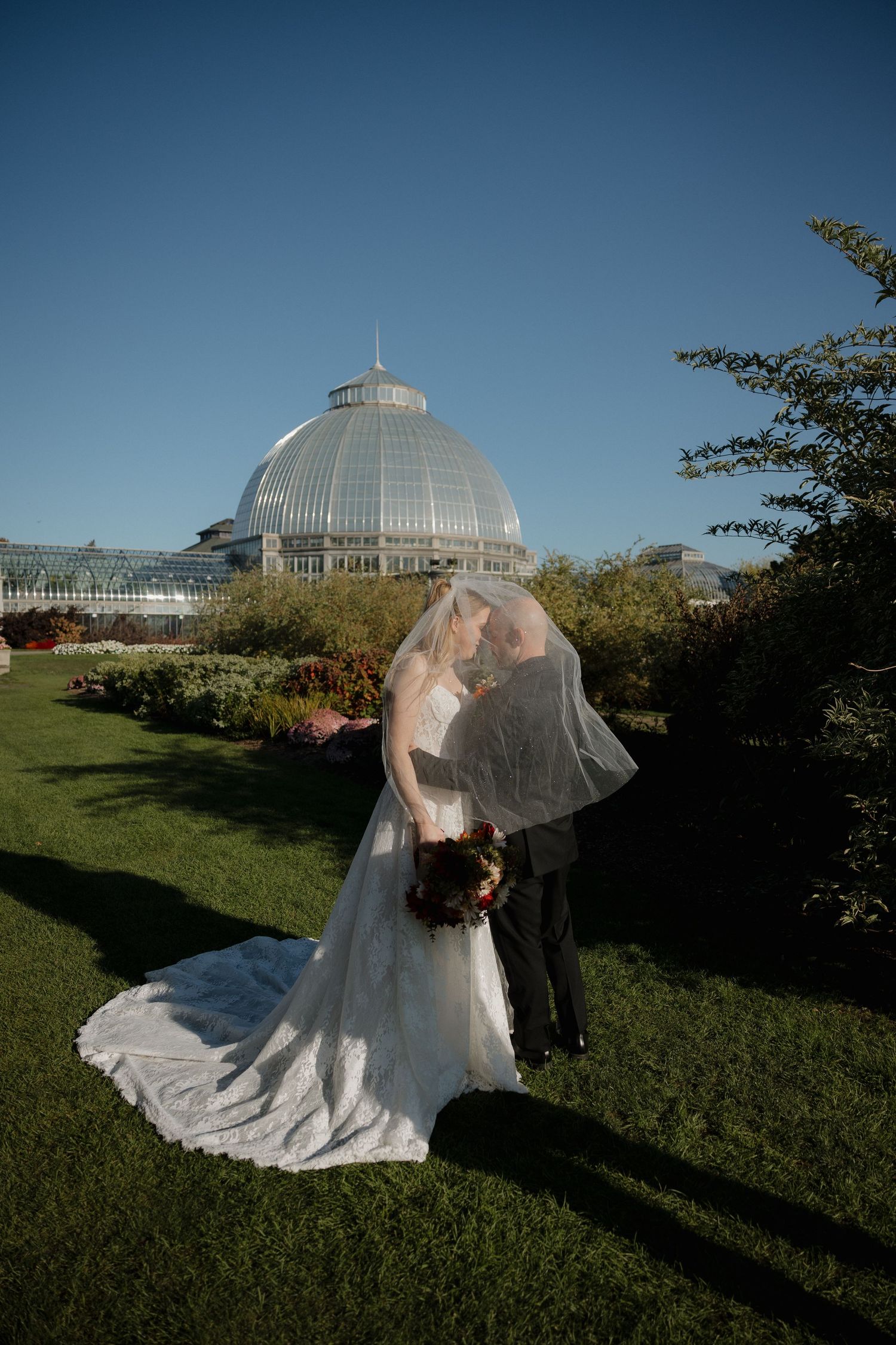 Bride and groom kissing under veil at Belle Isle Conservatory greenhouse dome on sunny wedding day.
