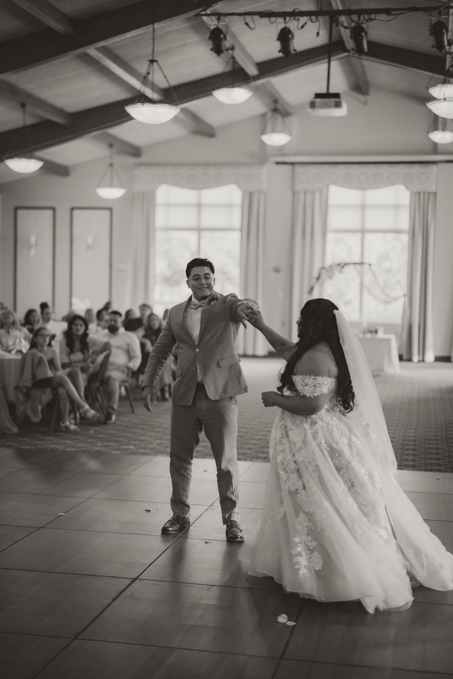 A couple shares their first dance in a bright reception hall filled with guests while the bride wears a flowing lace gown.
