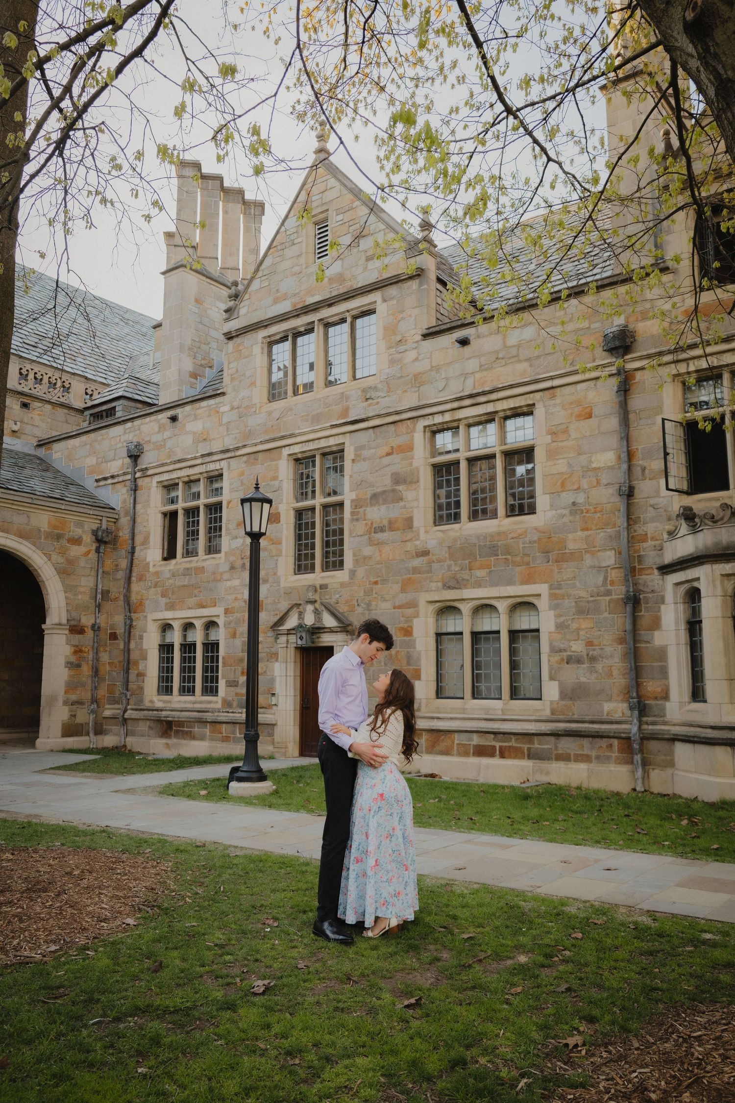 Couple embracing romantically in front of a historic stone building on a university campus in spring.