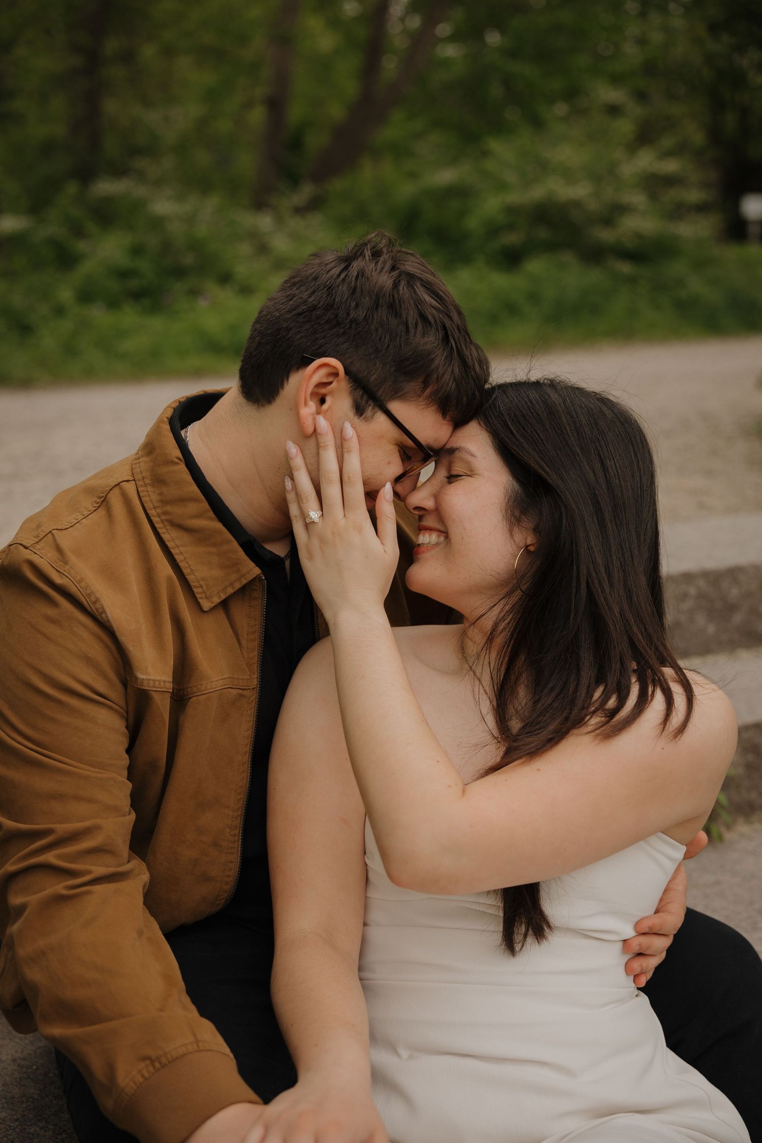 A couple sharing a romantic moment outdoors, smiling and touching foreheads, woman in white dress, man in brown jacket.
