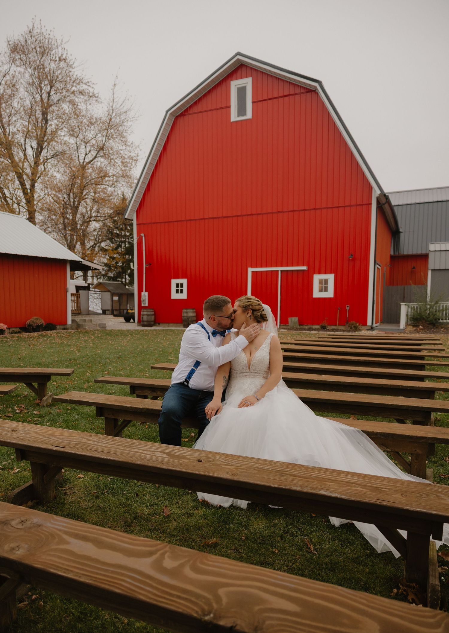 Bride and groom share a romantic kiss among wooden benches in front of a classic red barn at an outdoor fall wedding.