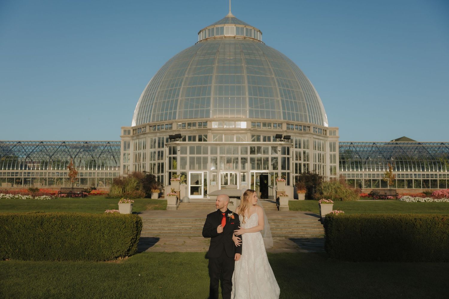 Bride and groom toasting in front of a grand glass dome greenhouse conservatory on a clear sunny day.