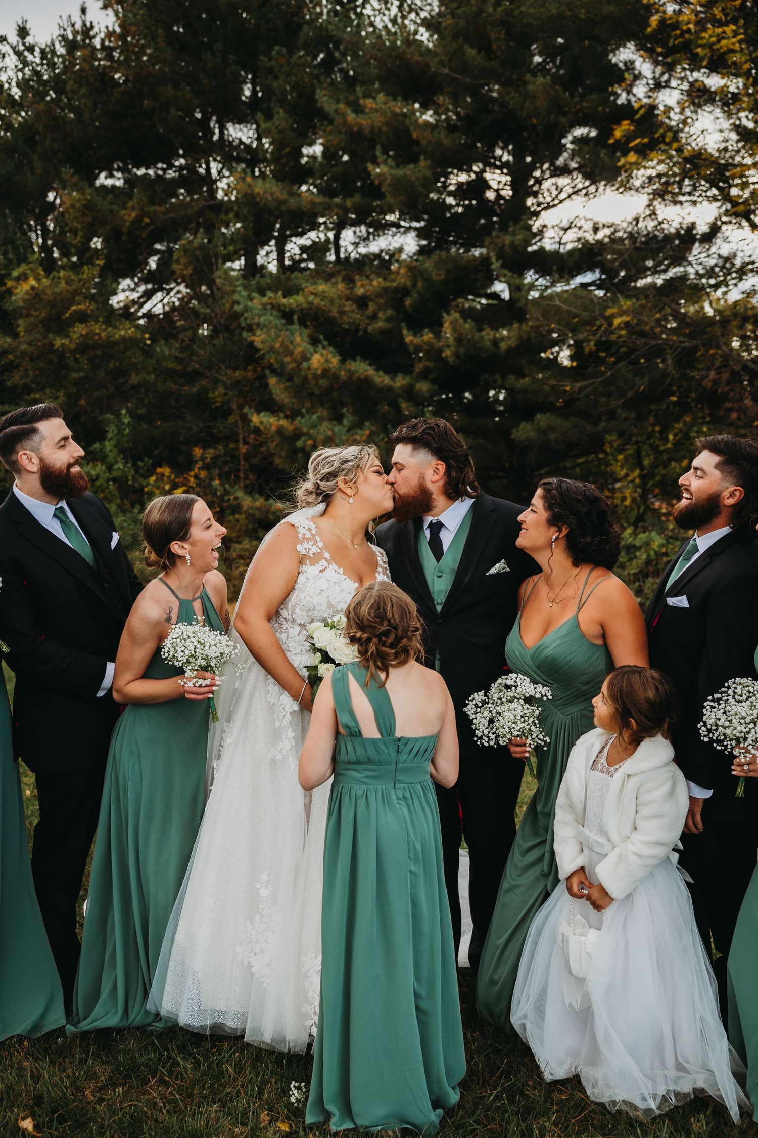 Wedding party in teal dresses and black suits laughing together outdoors with pine trees in background.