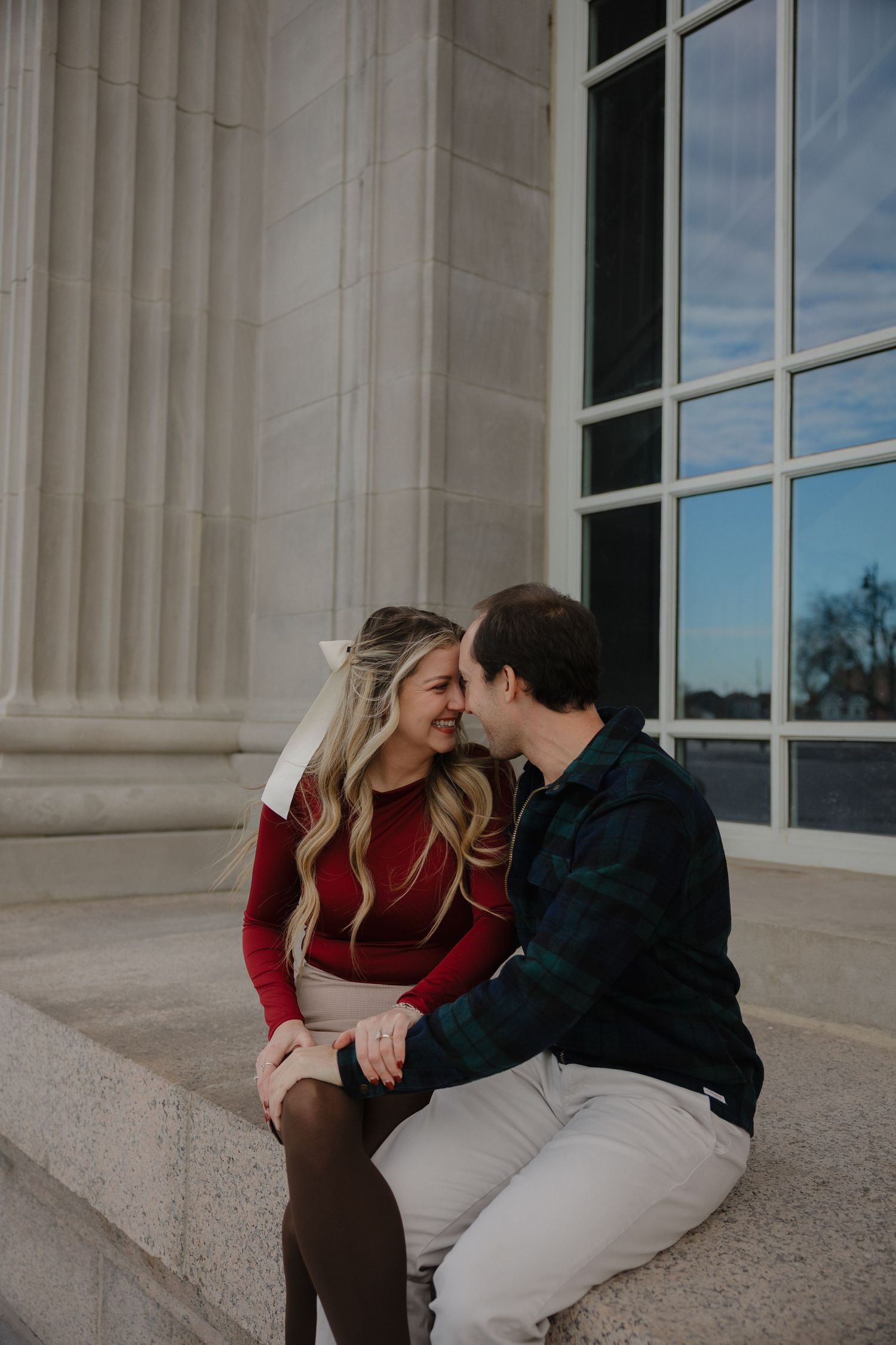 Couple sharing a romantic moment on stone steps near elegant columns and large windows outdoors.
