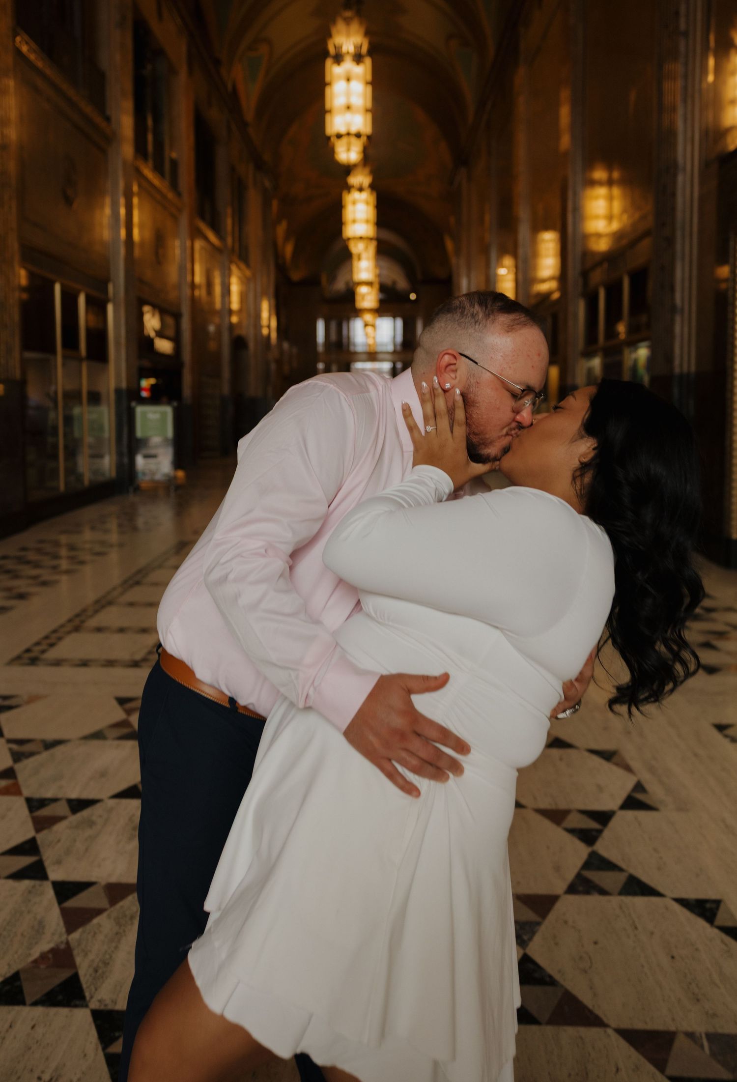 Couple sharing a romantic dip kiss in an elegant marble hallway with ornate chandeliers, bride in white dress.