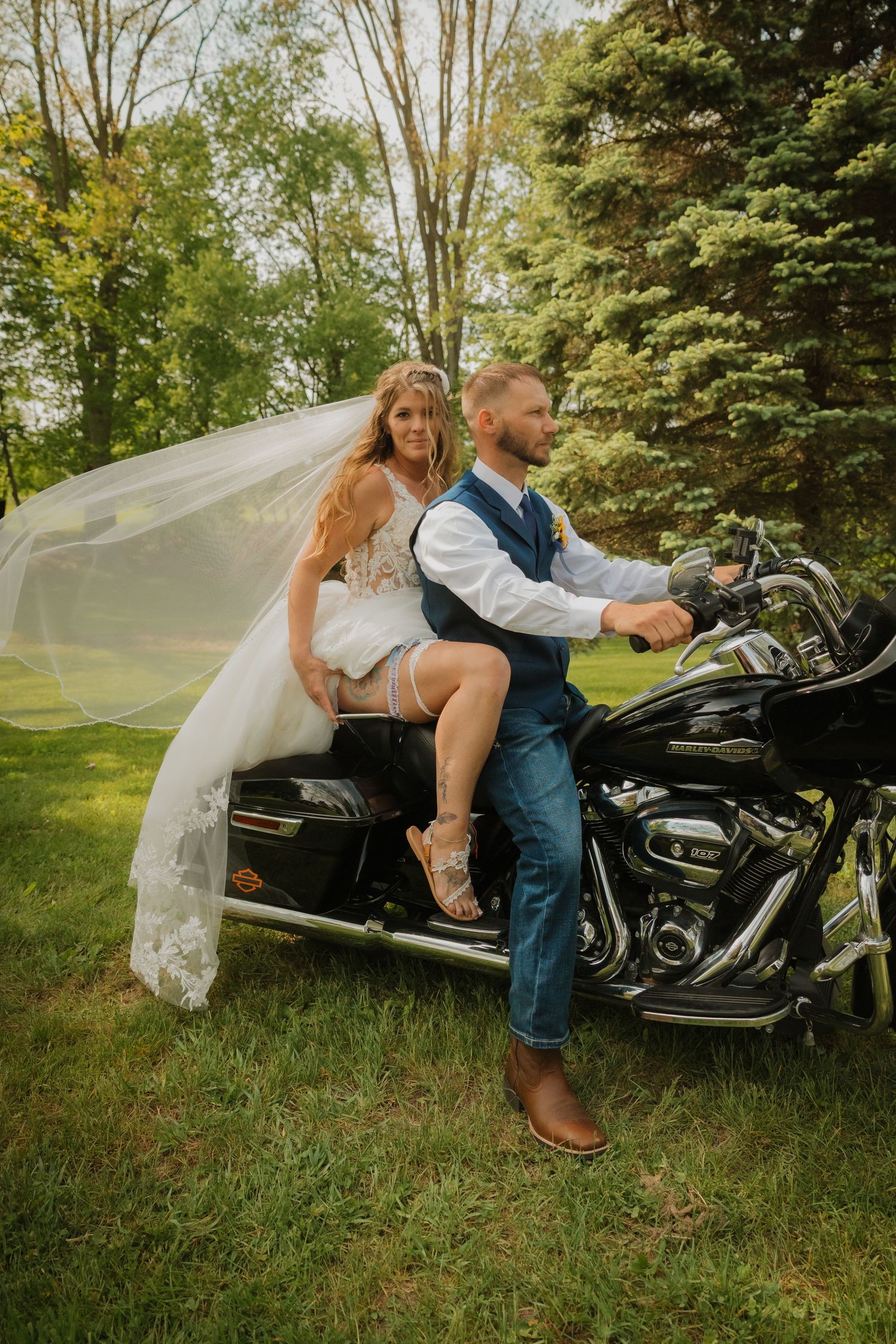 Bride and groom posing on a Harley-Davidson motorcycle outdoors, with her veil flowing in the wind amid lush green trees.