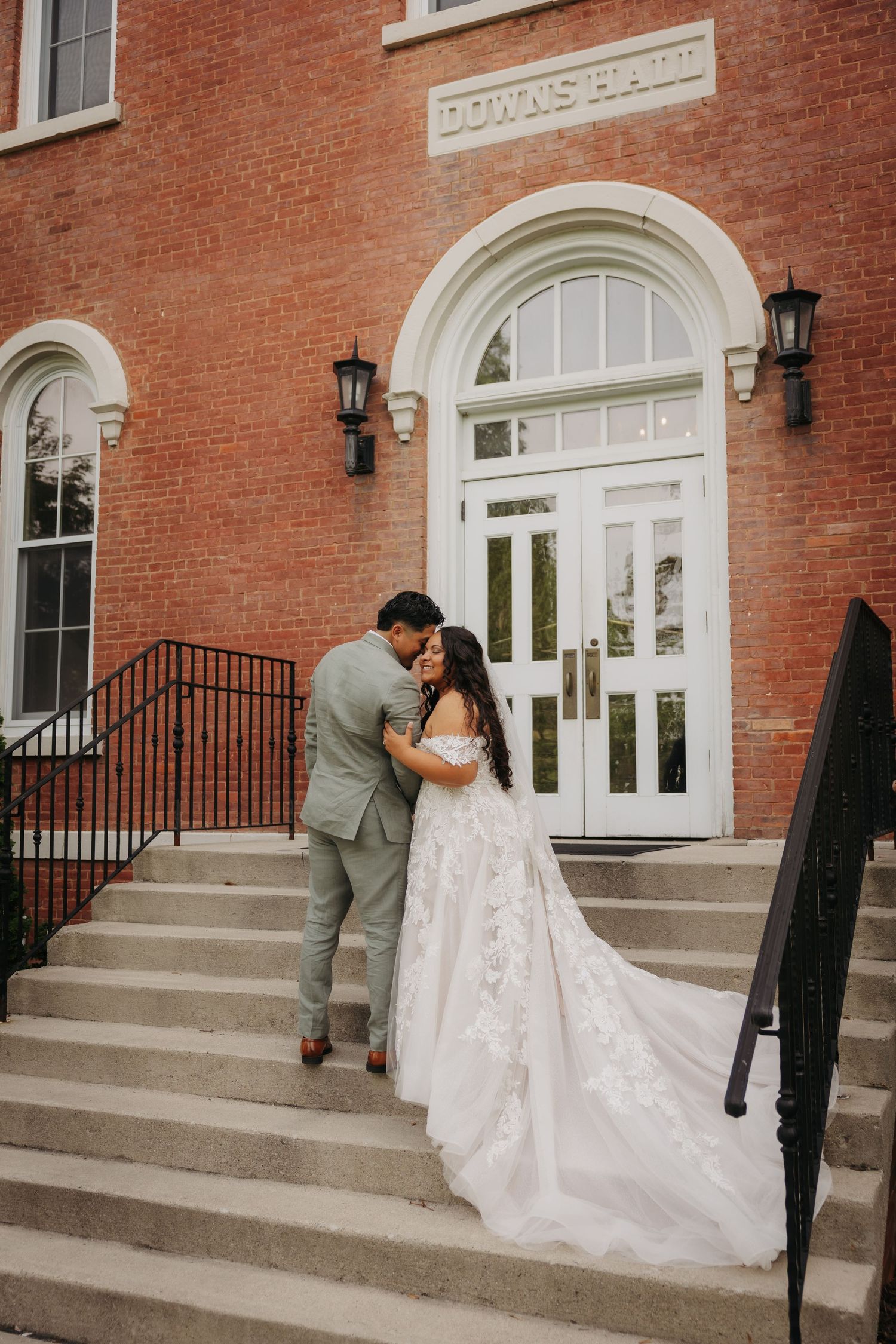 A bride and groom share an intimate moment on the steps of a historic red brick building with white trim and lanterns.