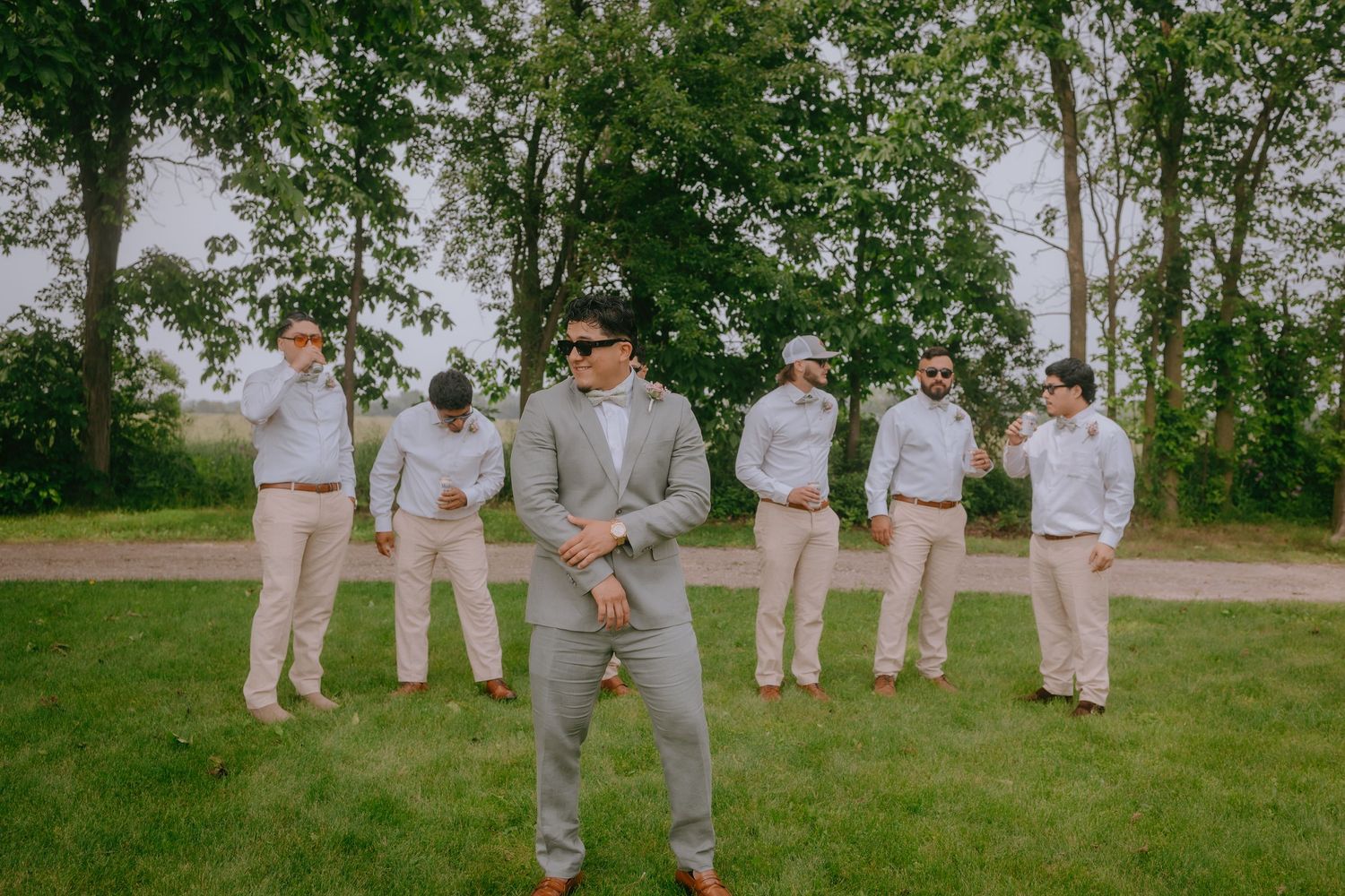 A wedding party stands on grass with trees, featuring groomsmen in light-colored suits arranged behind central figure.