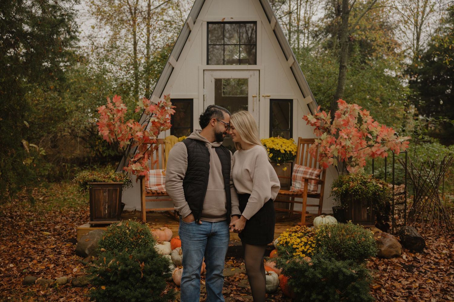 A couple stands together in a fall garden with orange leaves and rustic wooden buildings in the background.