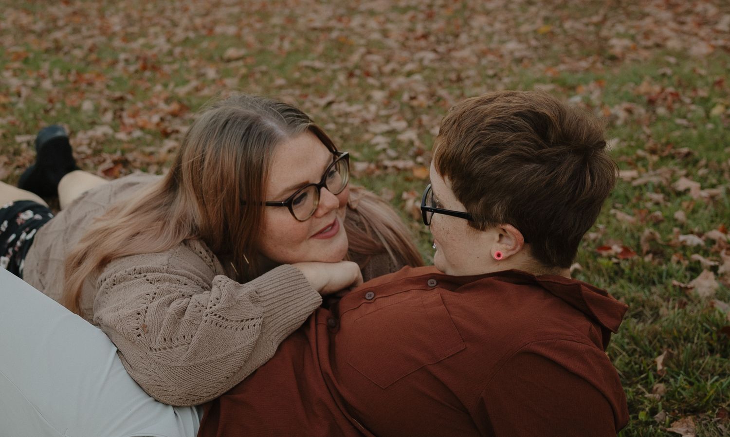 Couple with glasses lying on autumn leaves, smiling at each other in a romantic outdoor setting.