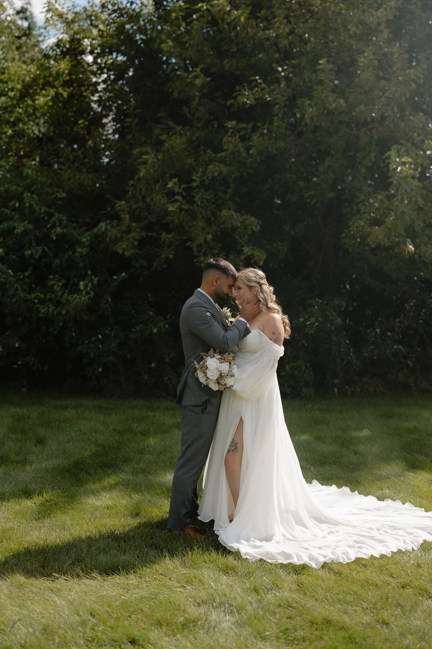 Couple in wedding attire embrace on sunlit grass with long white train flowing behind.