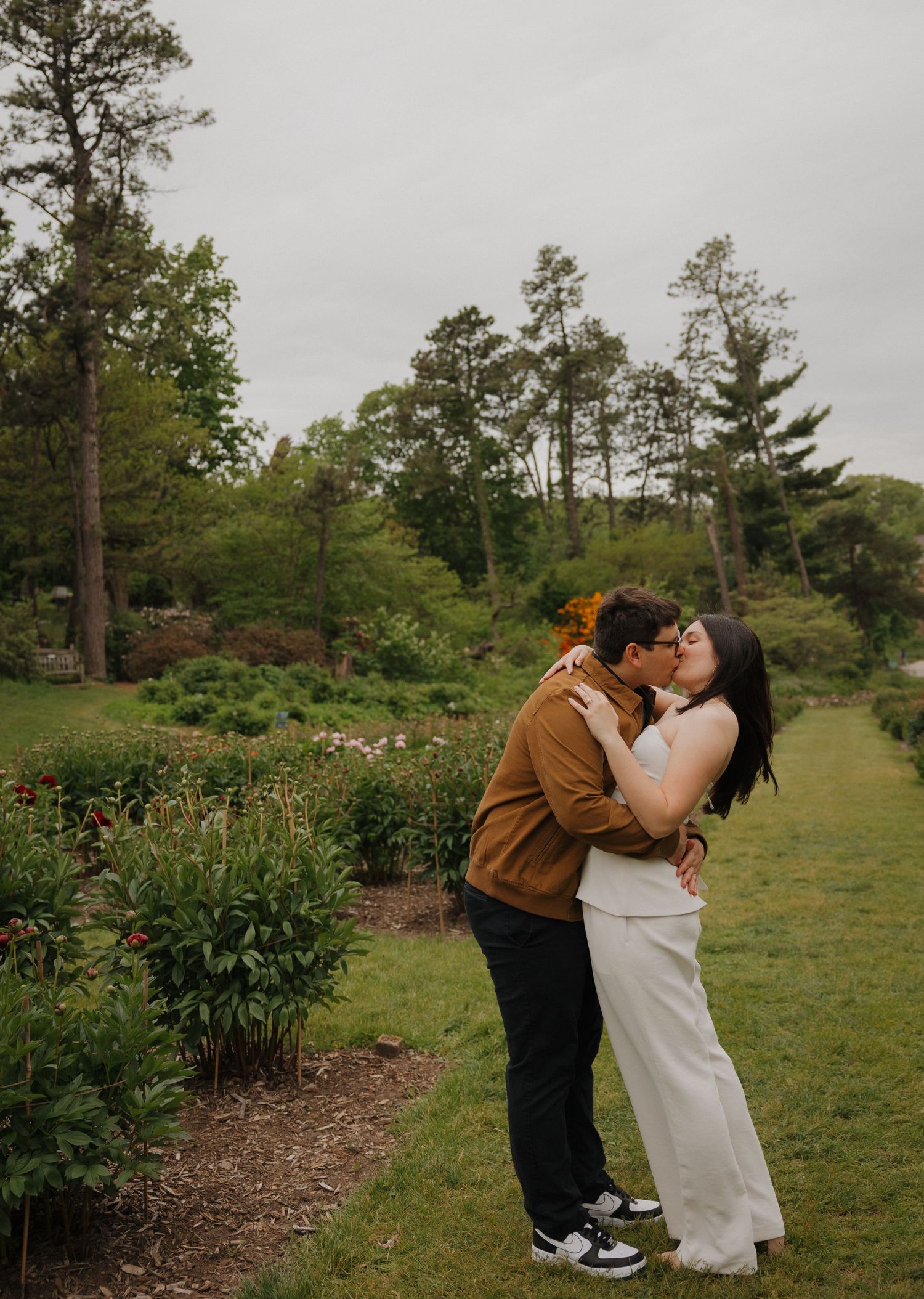 Couple sharing a romantic kiss in a lush garden setting, woman in white outfit, man in brown jacket.