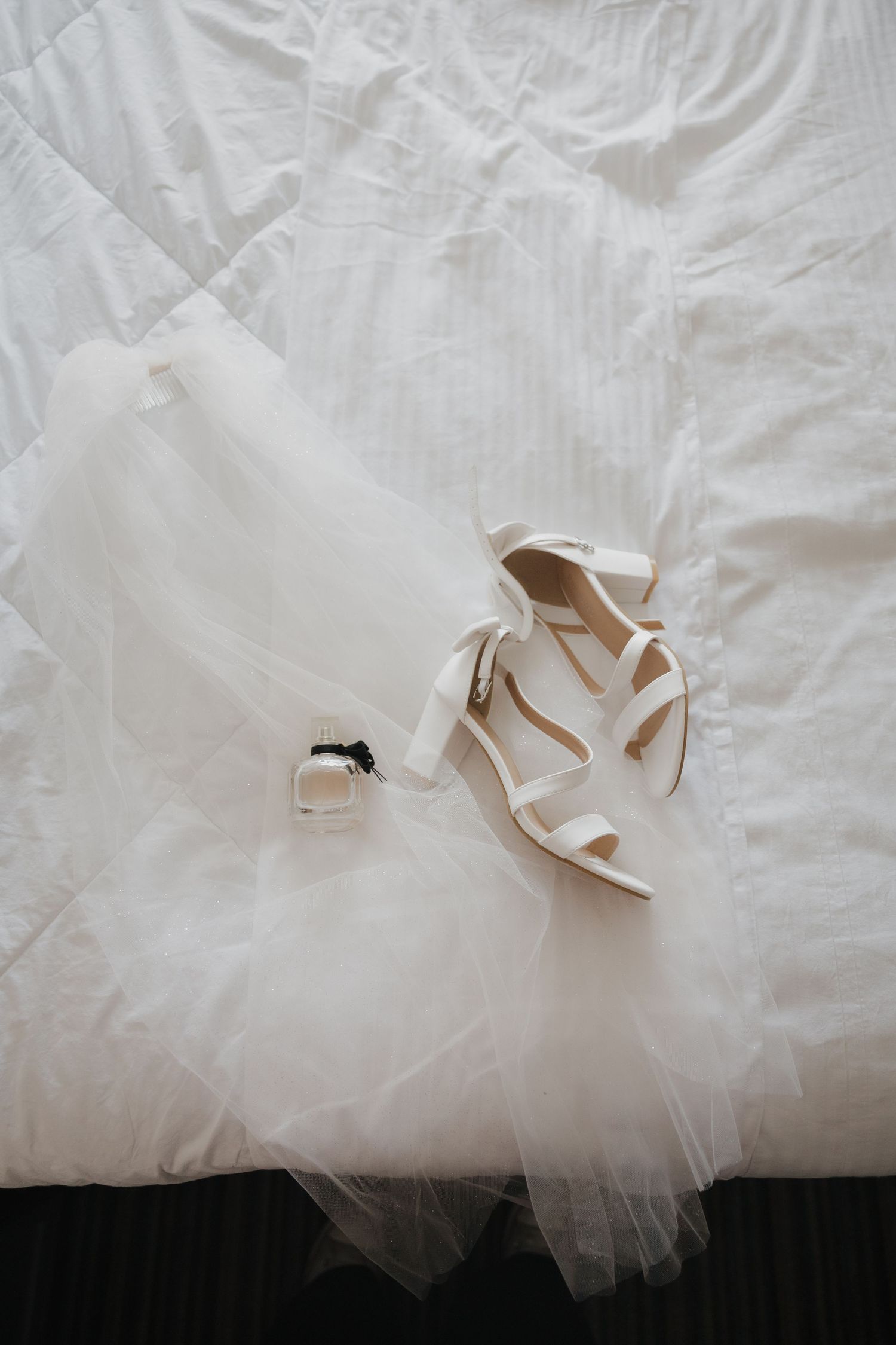 White bridal heels with ankle ties resting on a tulle veil beside a perfume bottle on a white bed.