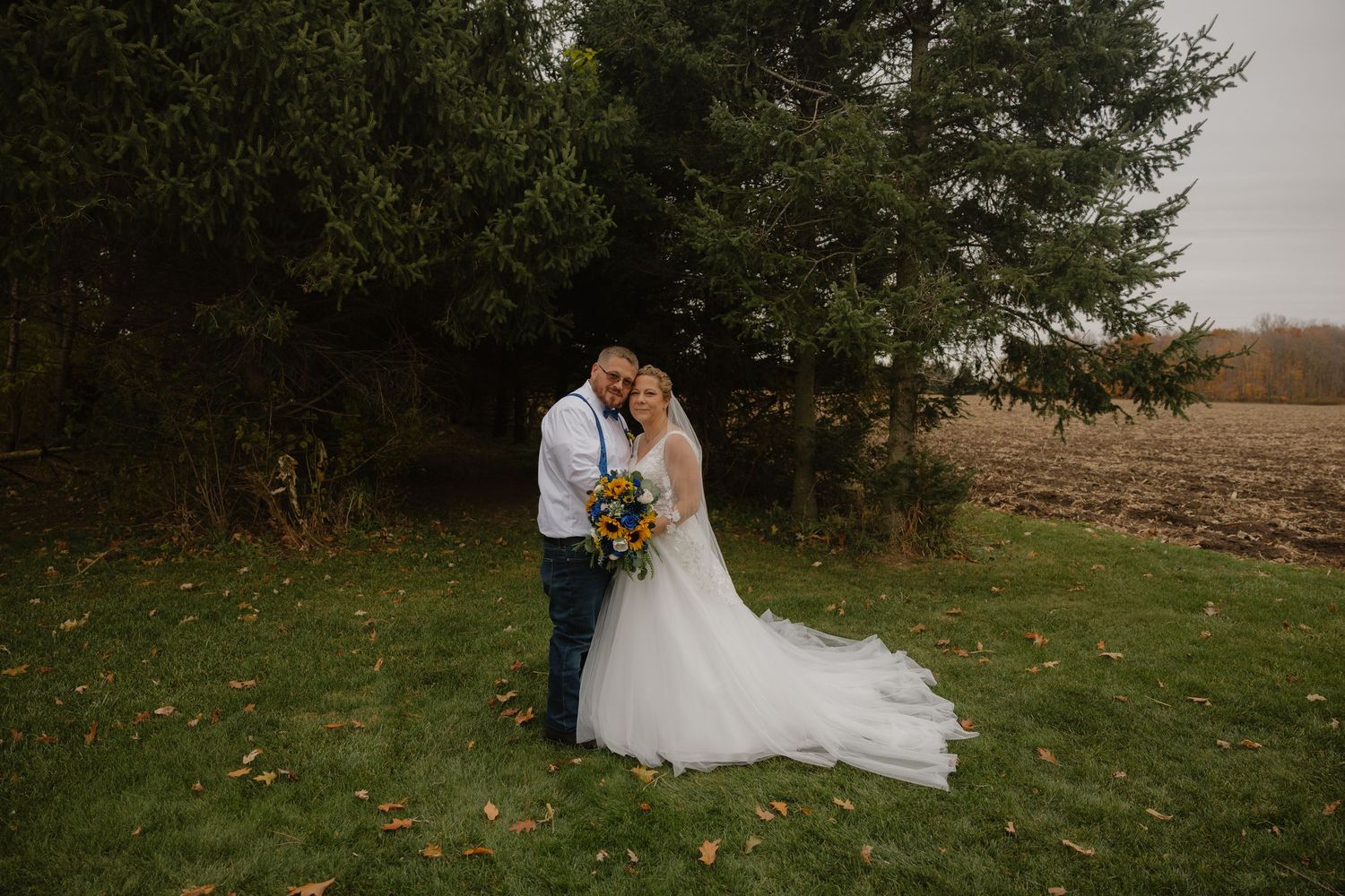 Bride and groom embracing outdoors near evergreen trees with autumn field backdrop, bride holding colorful bouquet.