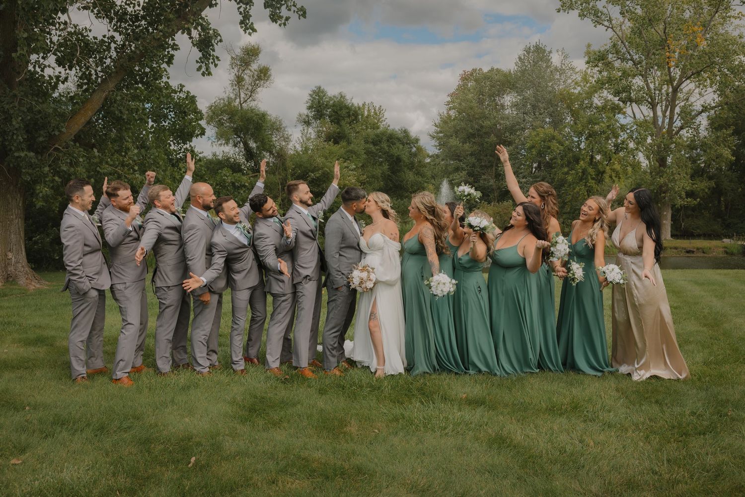 Full wedding party in mint green dresses and gray suits posed on grassy hill for group photo.