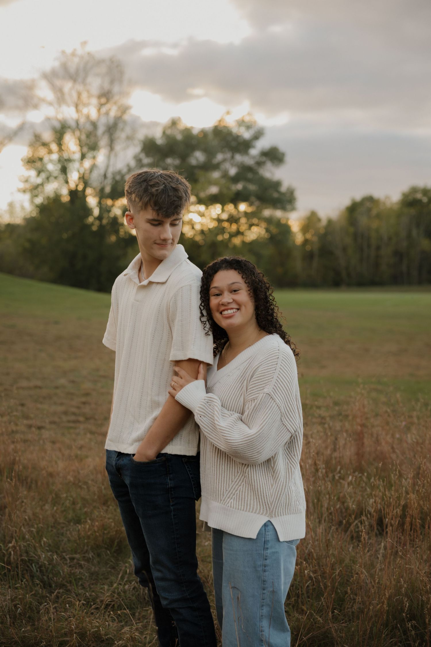 A couple in casual white sweaters share a tender moment together in a sunlit autumn field at golden hour.