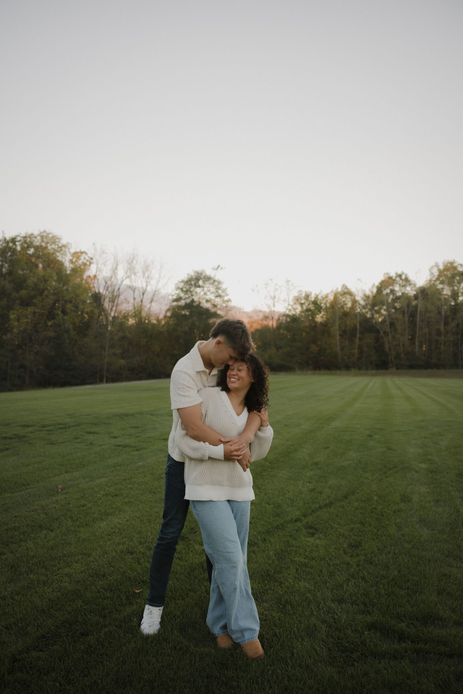 A couple embraces lovingly in a serene green field at sunset with trees in the background.