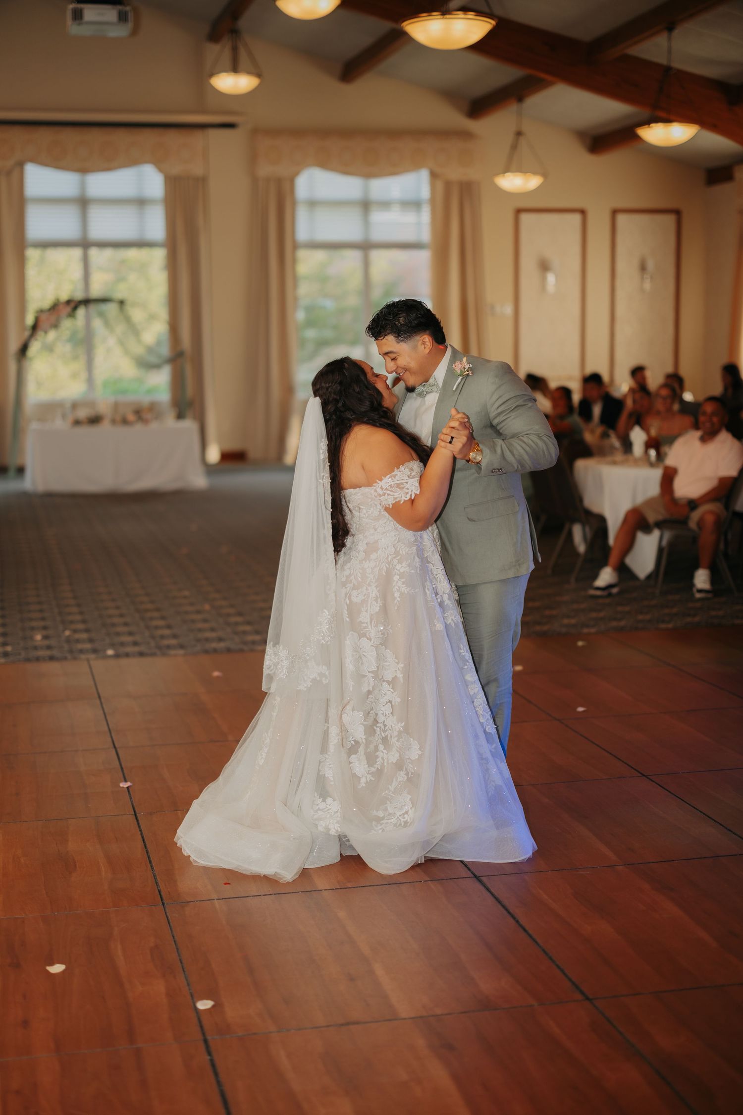 A couple shares their first dance as newlyweds on a wooden dance floor in a sunlit reception venue with exposed beams.