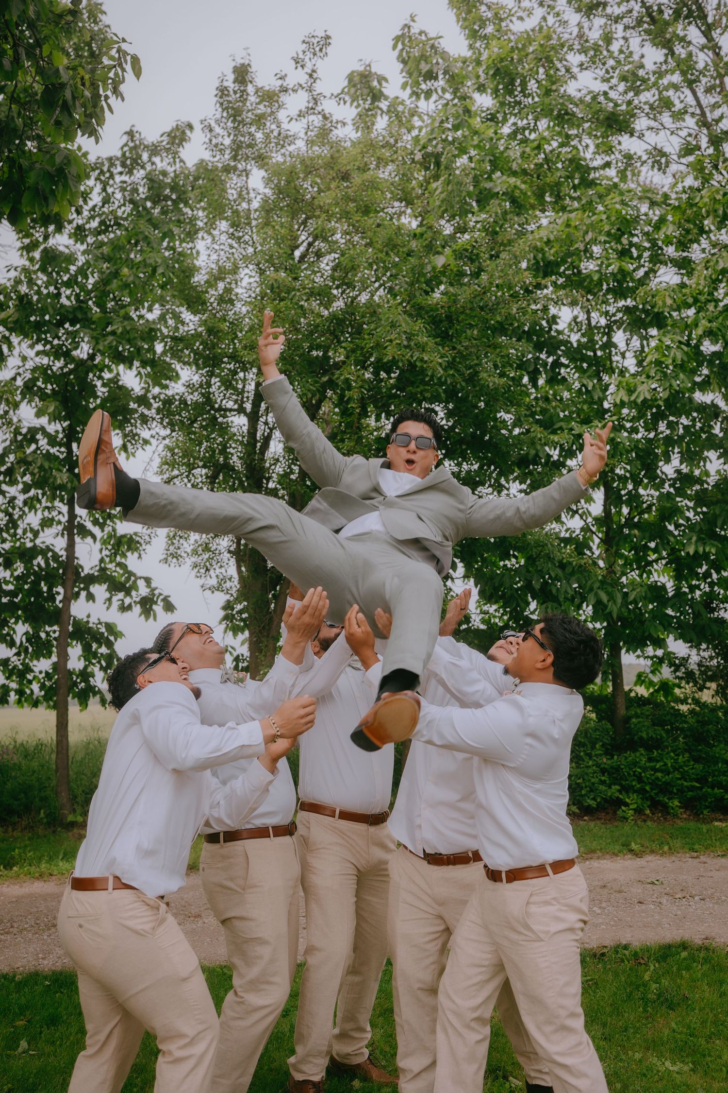 Groomsmen in light khaki pants and white shirts toss and catch someone in a grey suit during outdoor wedding photo.