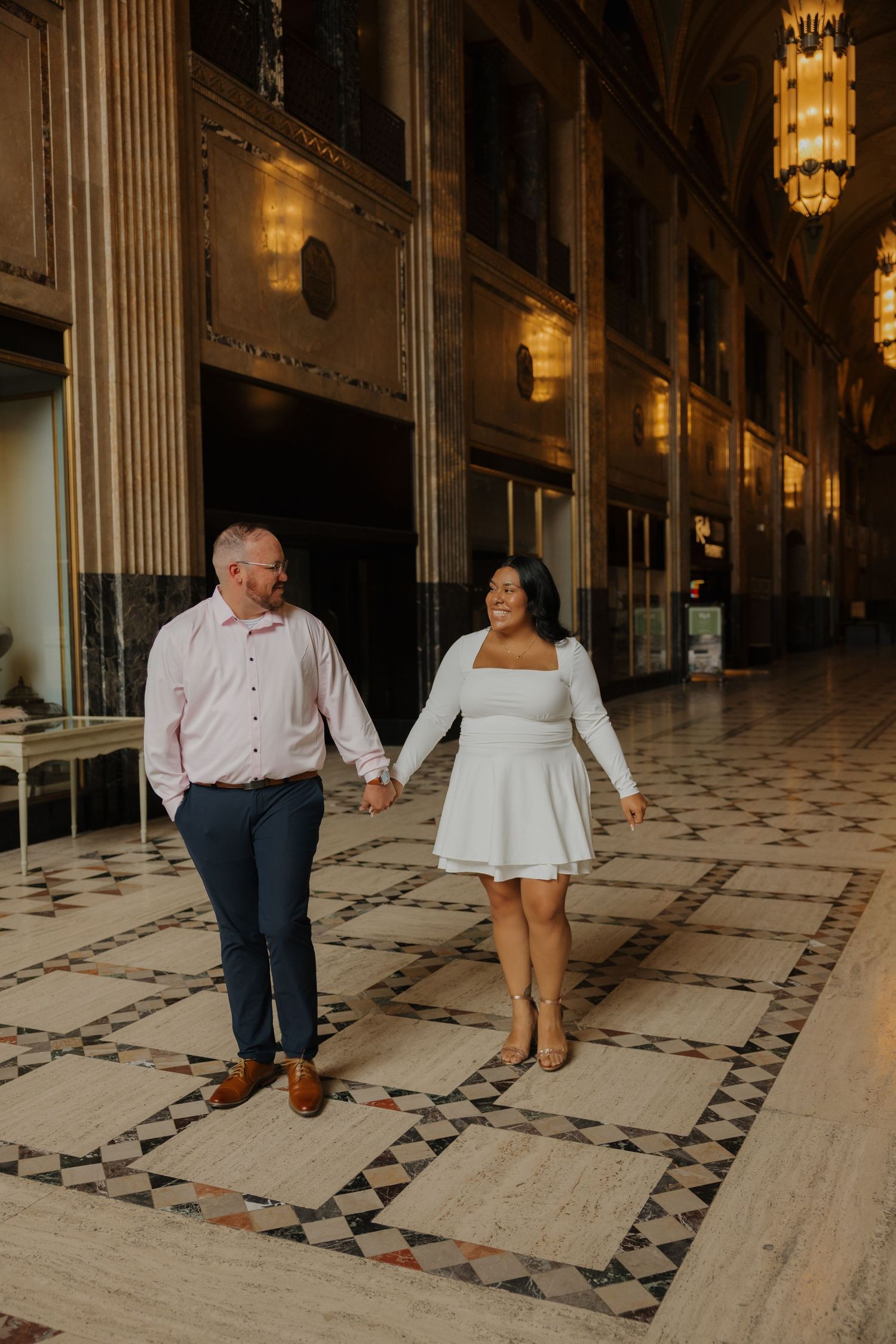 Couple holding hands walking through elegant marble hallway with ornate chandeliers during engagement session.