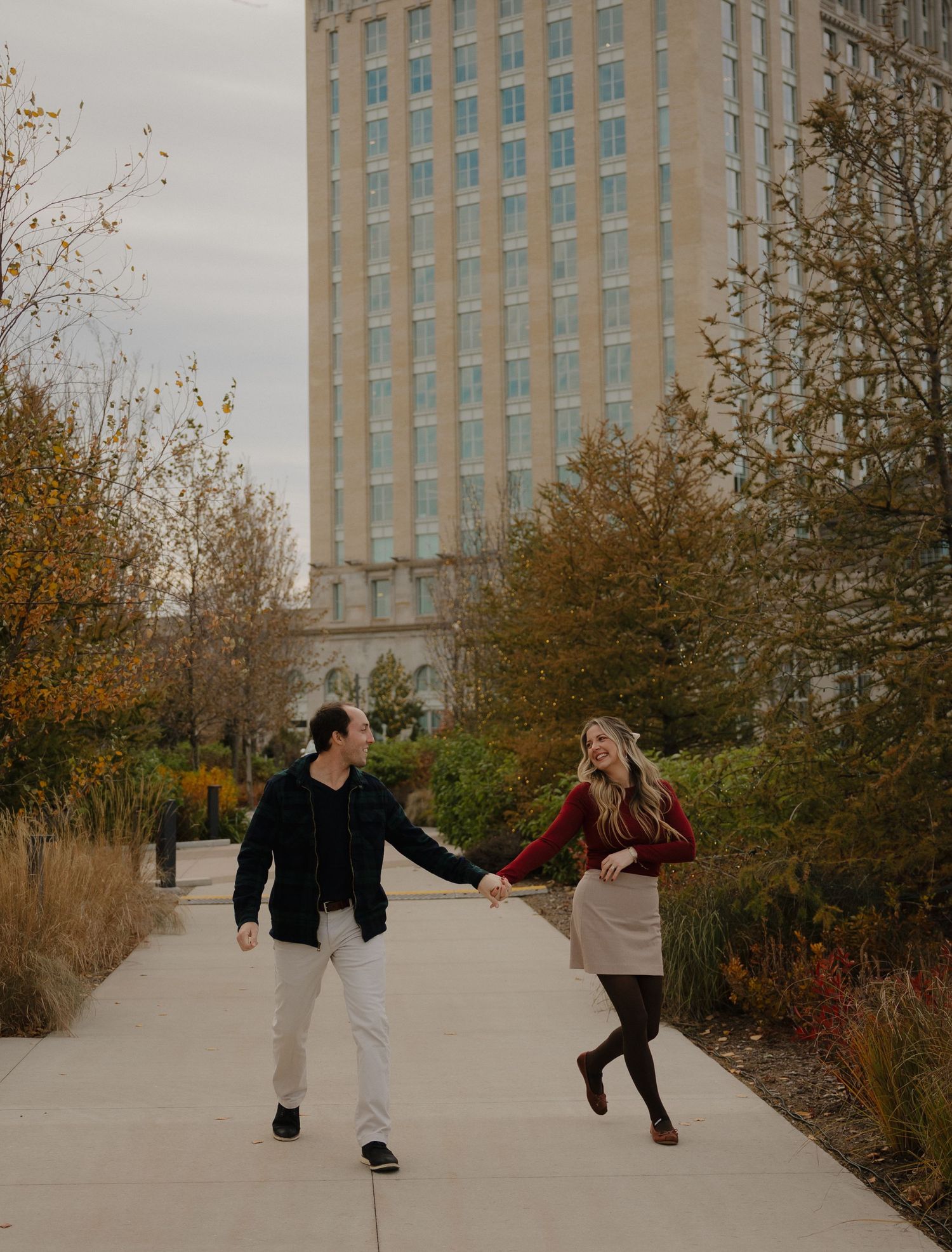 A couple holds hands and walks along an urban pathway surrounded by autumn trees and a tall building in the background.