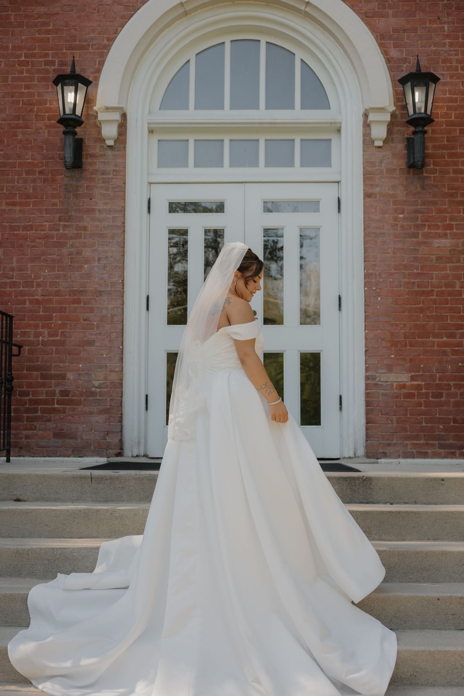 Bride in off-shoulder white ball gown with cathedral veil stands on brick church steps, showing elegant train.