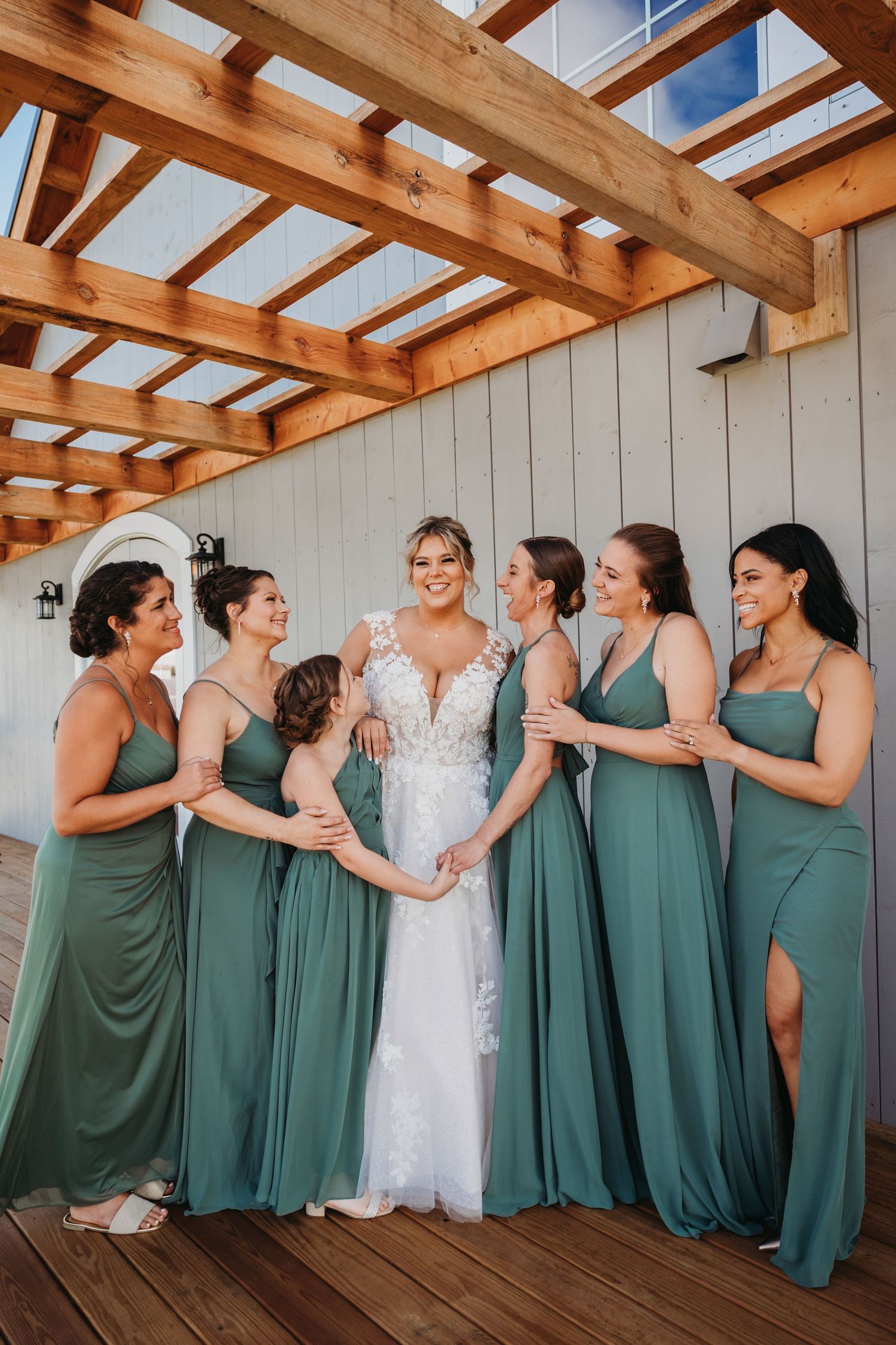 A bridal party in matching sage green dresses poses together on a wooden deck.