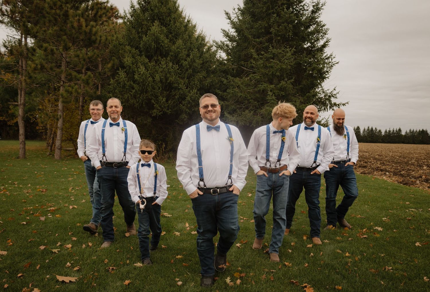 Groomsmen in white shirts, blue bow ties, and suspenders with jeans walk outdoors near evergreen trees at a wedding.
