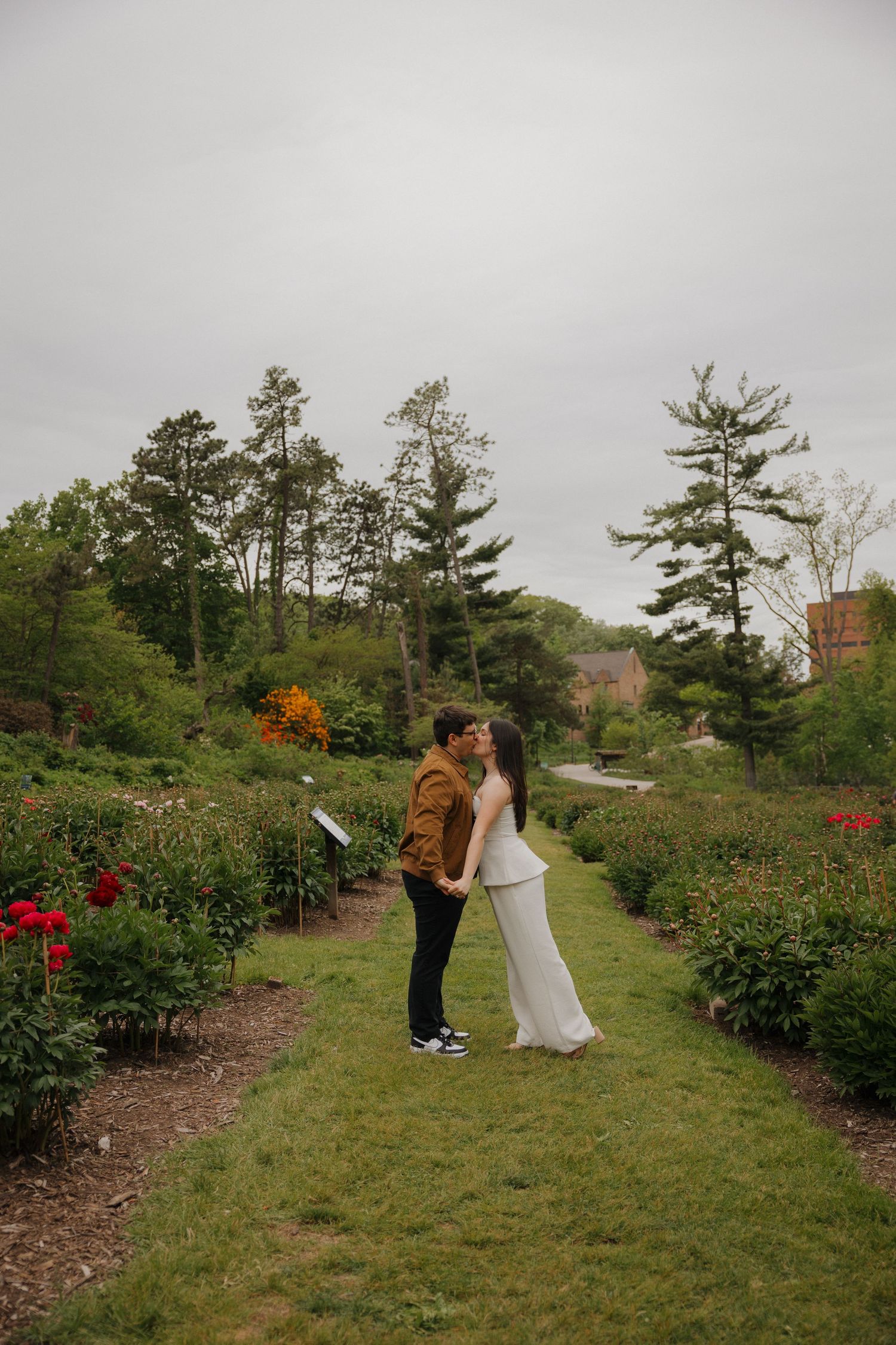 A couple shares a romantic kiss in a lush garden pathway surrounded by colorful flowers and tall trees on a cloudy day.