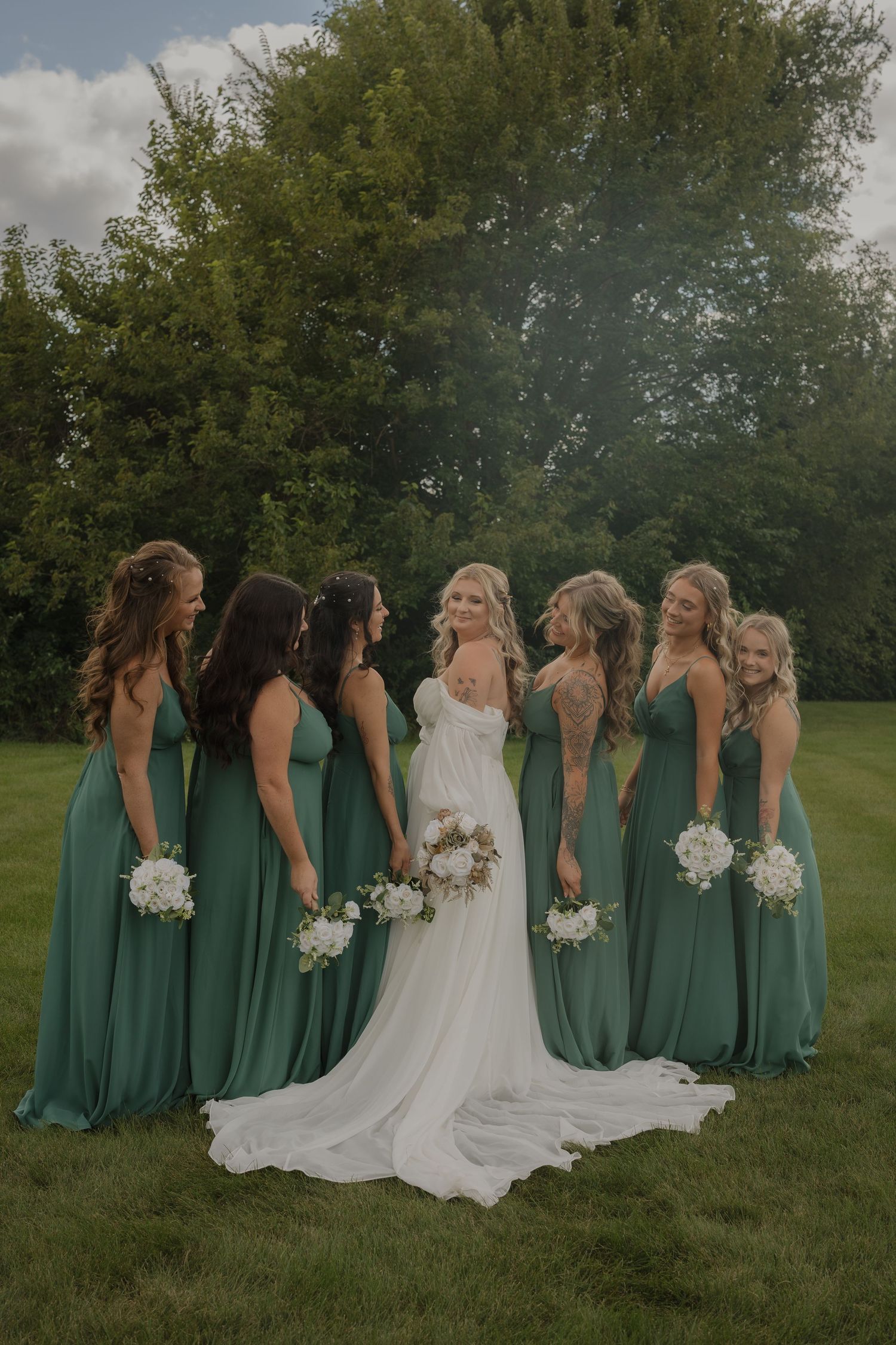 Bridesmaids in emerald green dresses stand with bride in white gown against natural outdoor backdrop.