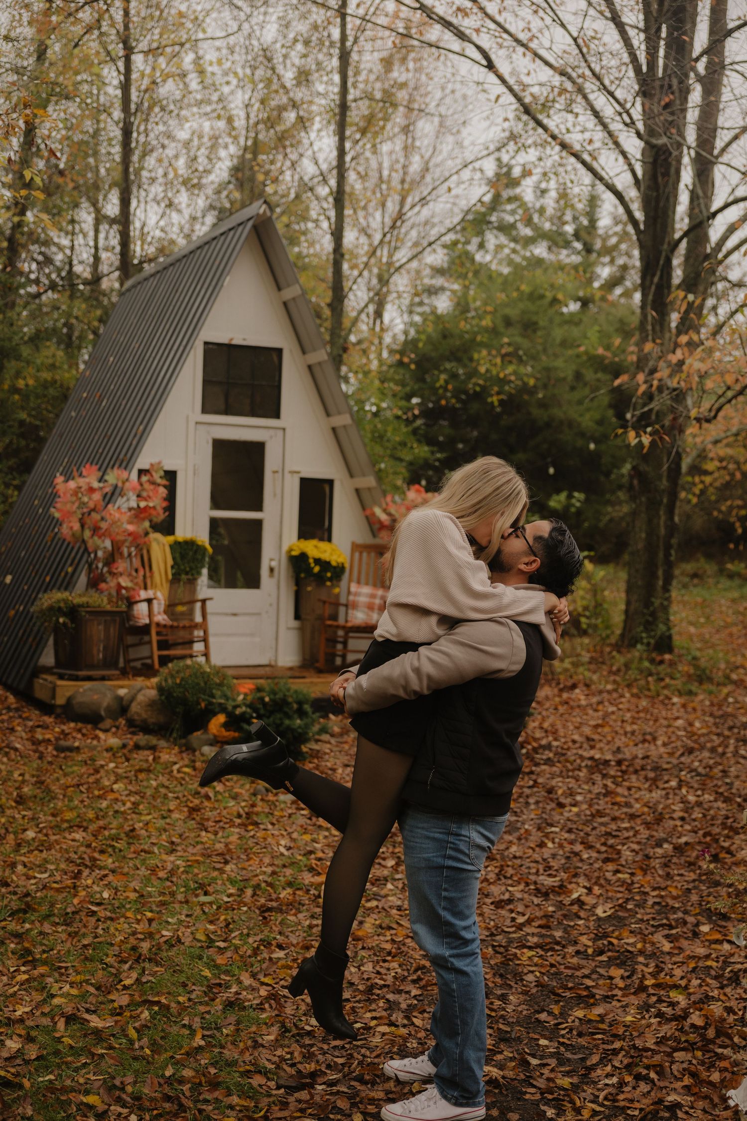 Two people share playful moments in front of a rustic A-frame cabin surrounded by autumn foliage.