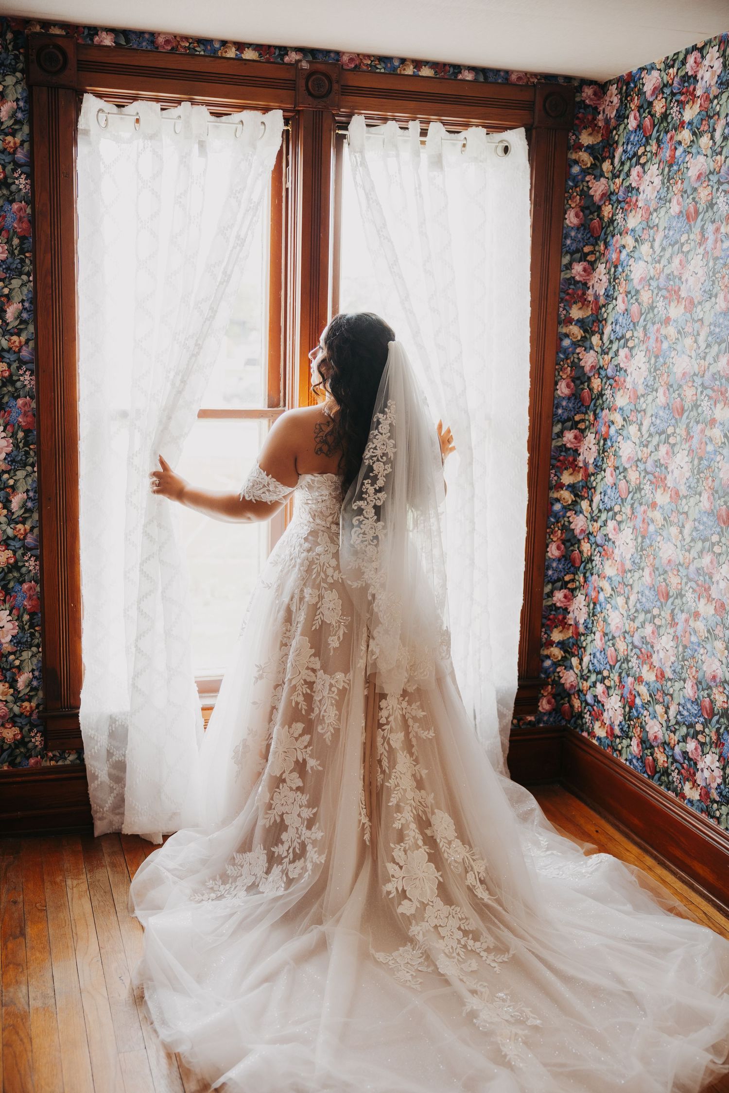 A bride in a flowing lace wedding gown and veil stands at a window with floral wallpaper in a vintage-style room.