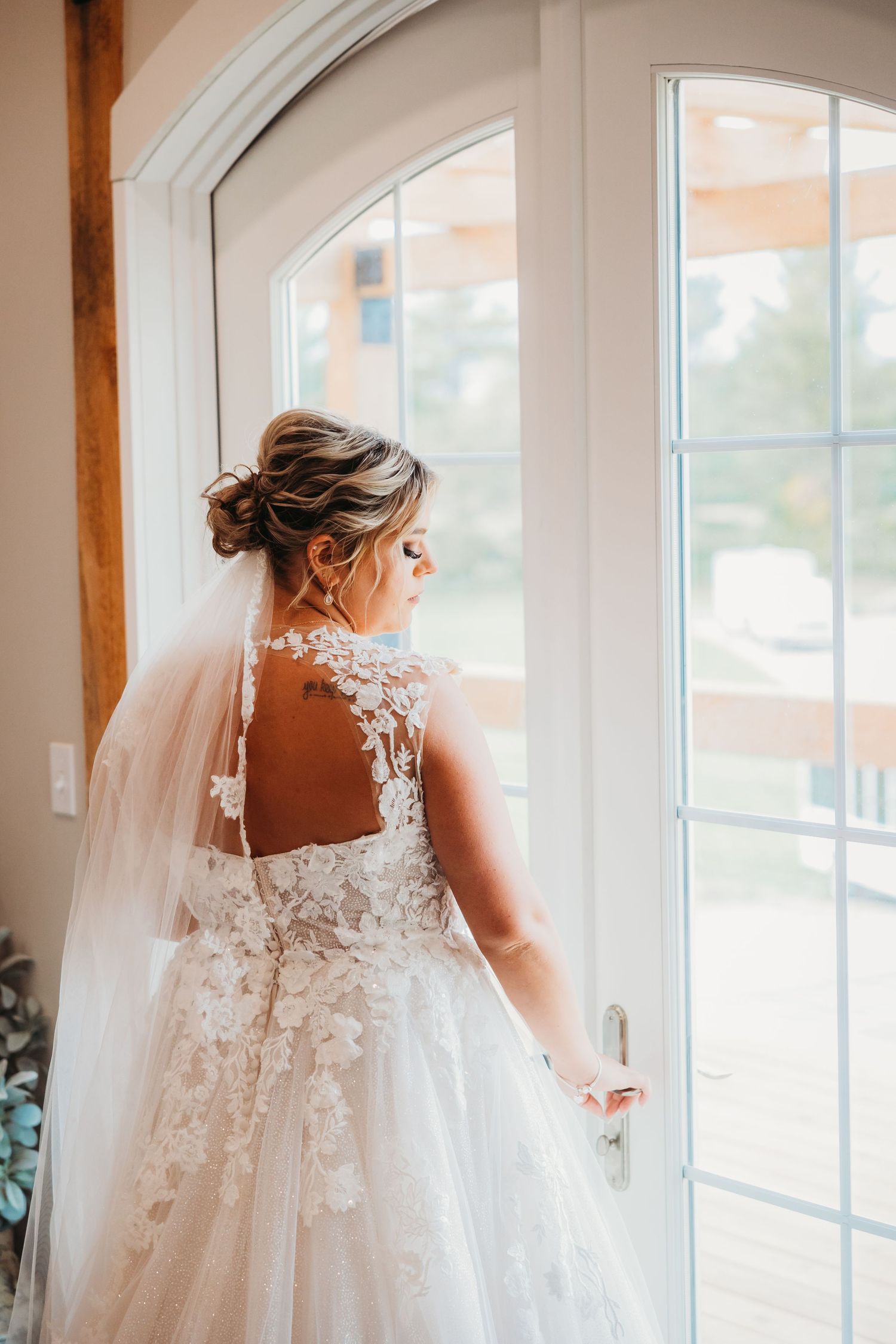 A bride in a lace wedding gown stands by a large arched window.