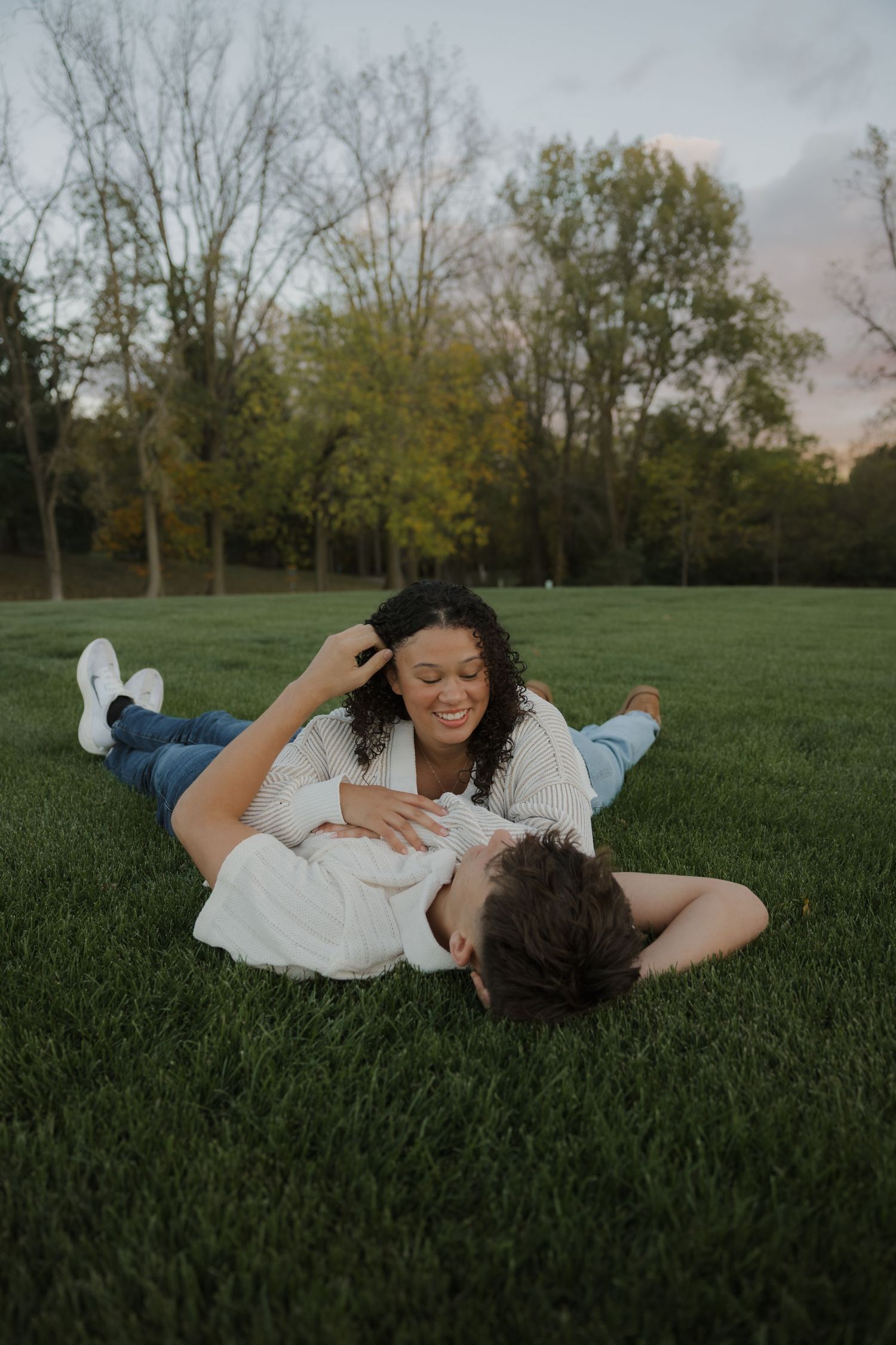 A couple lies together in the grass, sharing joyful laughter in a park setting.
