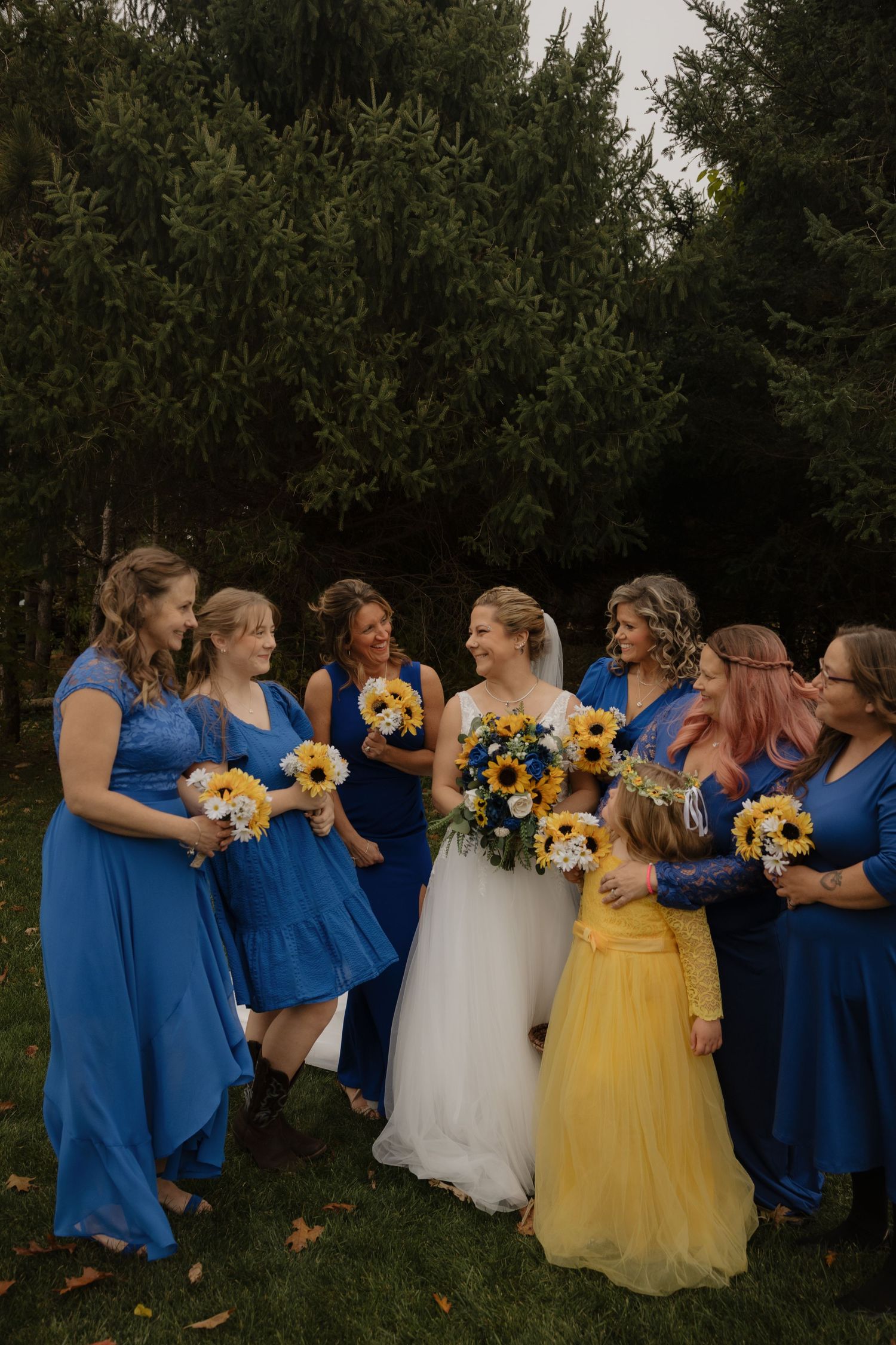Bride in white gown with bridesmaids in royal blue dresses and flower girl in yellow, holding sunflower bouquets outdoors.