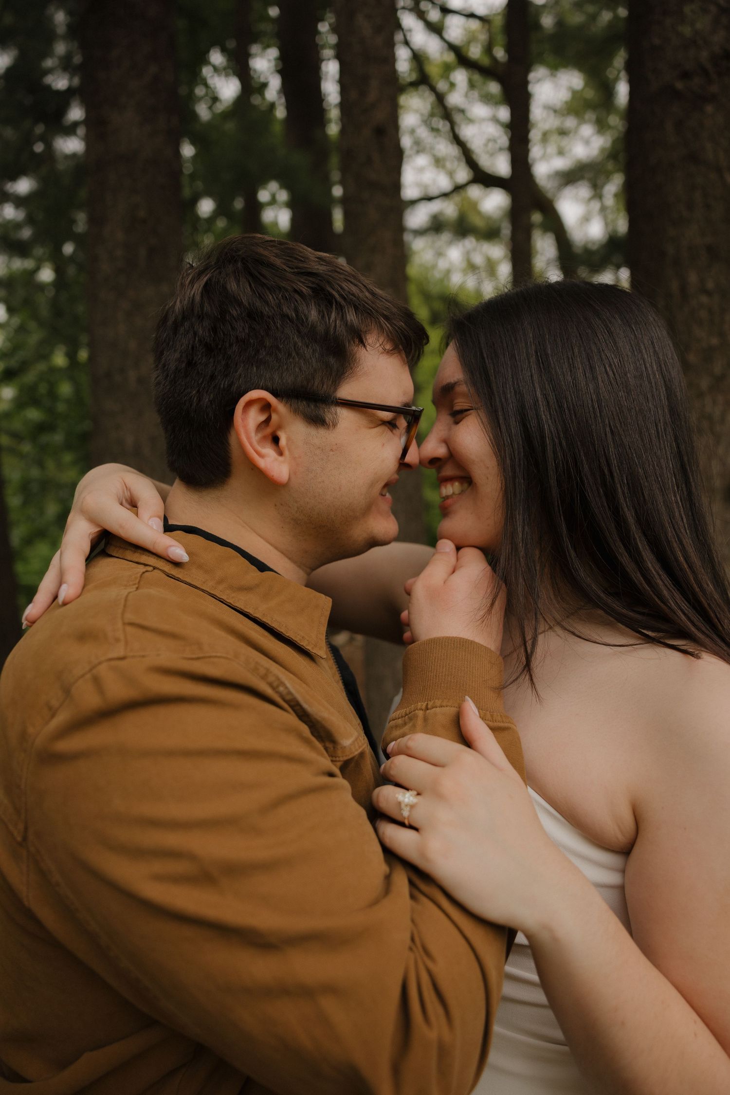 A couple shares a romantic moment in a forest, smiling and embracing, the man in a brown jacket and woman in white.
